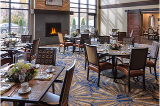 A dining room with several round and rectangular tables set with plates, cups, and floral centerpieces. The room features large windows letting in natural light, a lit fireplace, and a patterned carpet. Chairs are arranged around the tables, and there is a sliding barn door leading to another room.
