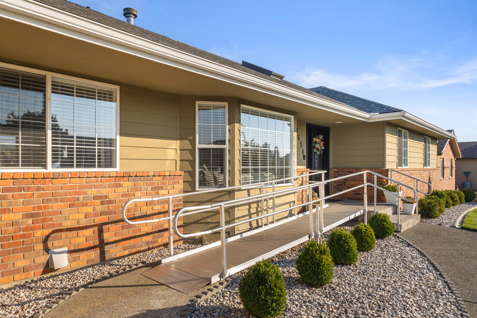 Ramp-accessible entrance to a single-story beige-brick senior home with shrubs.