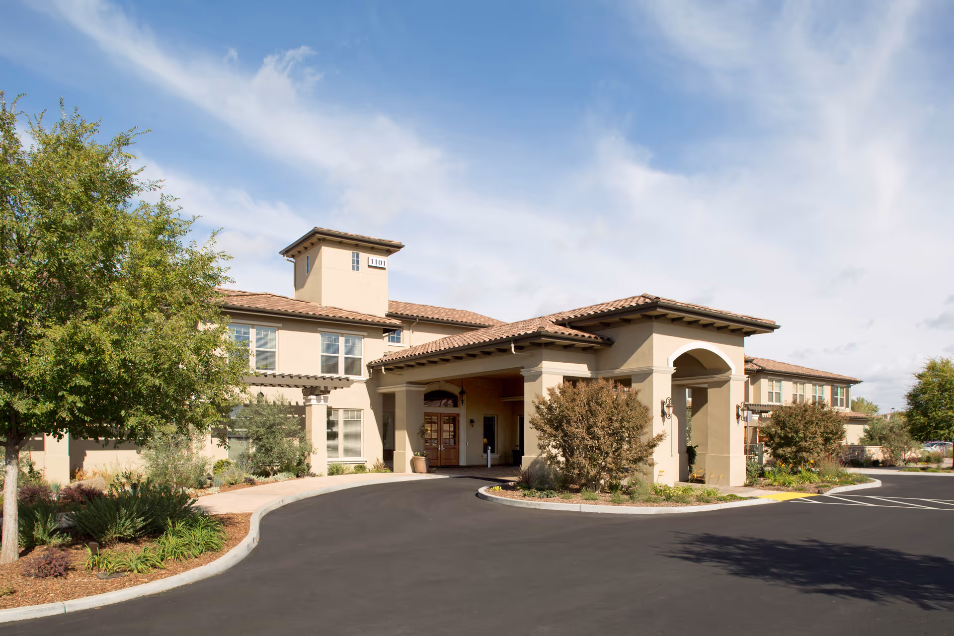 Exterior view of Oakmont of Roseville senior living facility showing a two-story beige building with a covered entrance, surrounded by landscaped greenery and a clear blue sky.