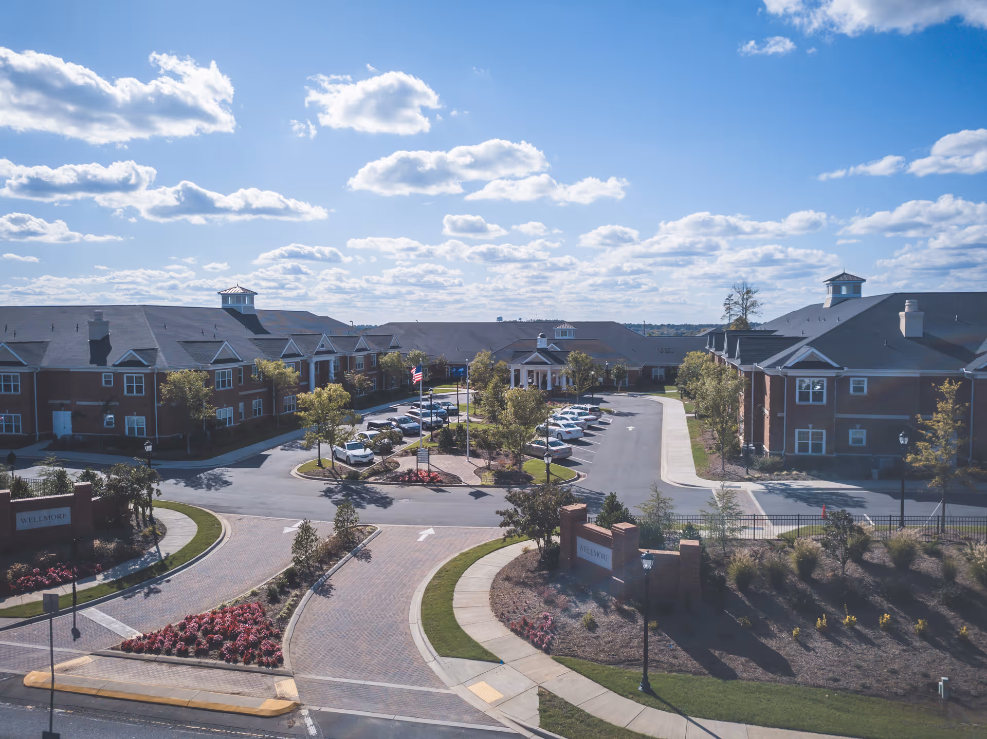 Aerial view of Wellmore of Tega Cay senior living facility showing multiple brick buildings with dark roofs surrounding a parking lot with several cars. The entrance features landscaped areas with flowers and trees under a partly cloudy blue sky.