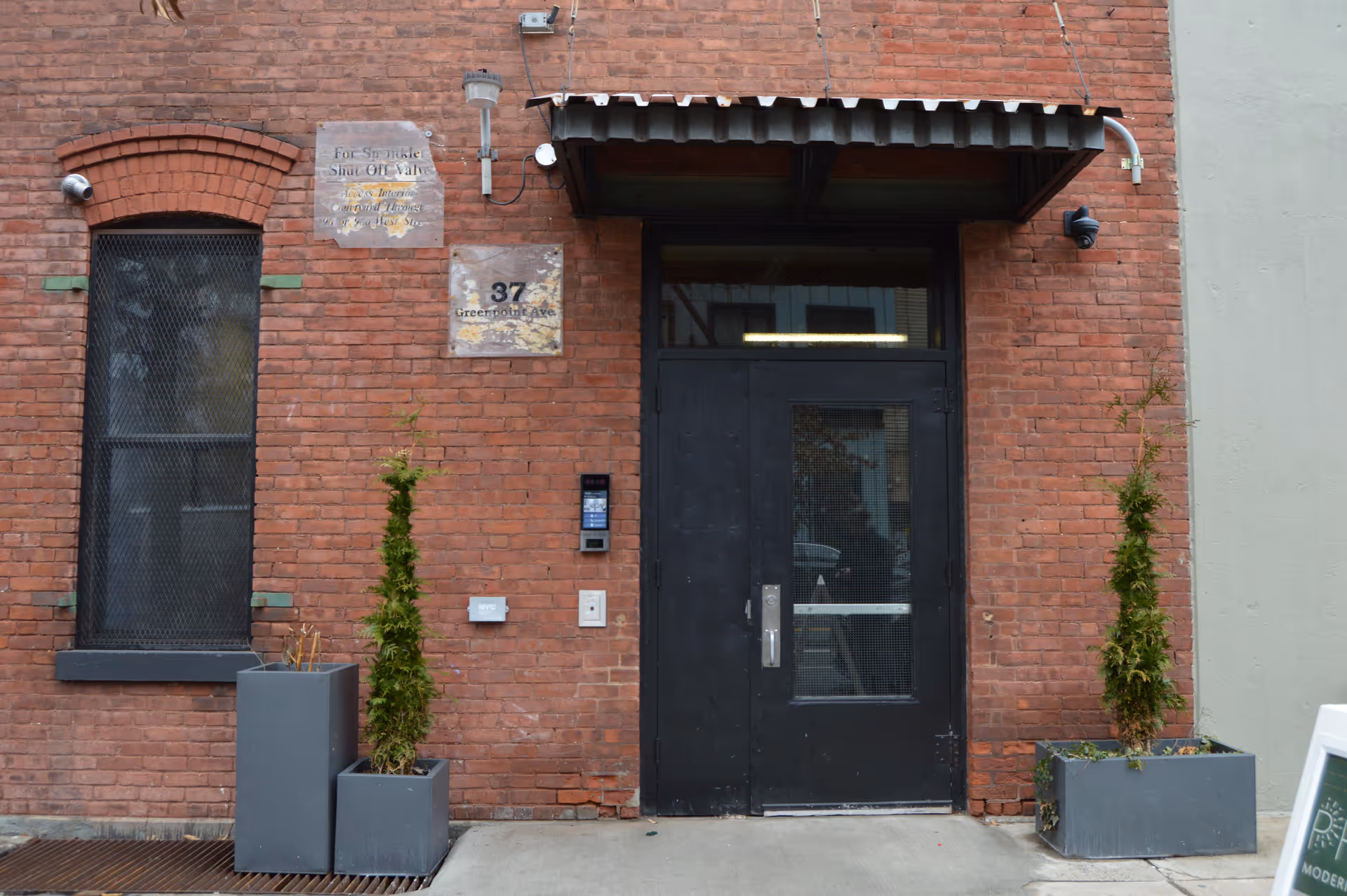 Brick building entrance with a black door, sign plaques, and tall potted trees.