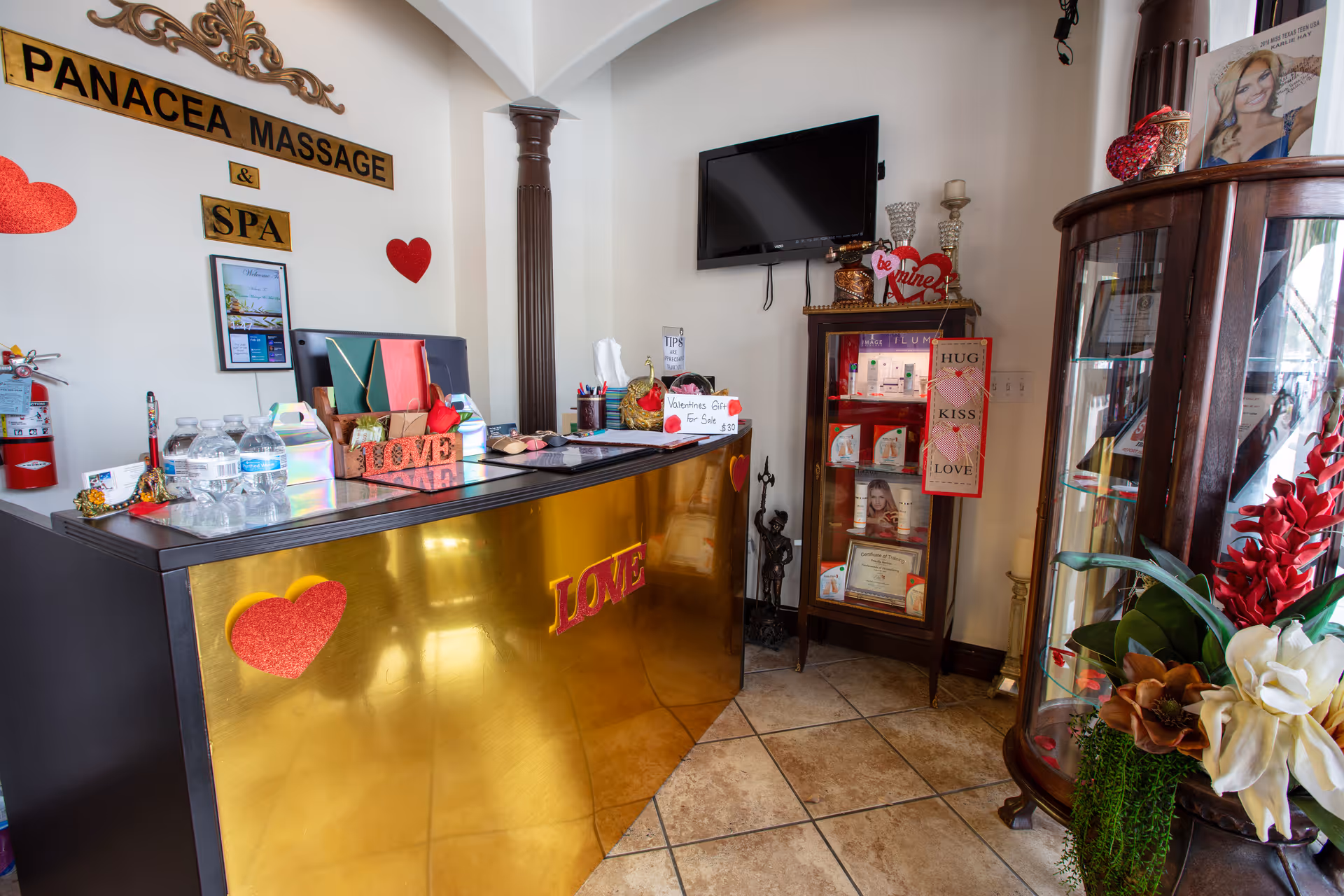 Reception area with a gold counter, Panacea Massage and Spa sign, TV, shelves, flowers.