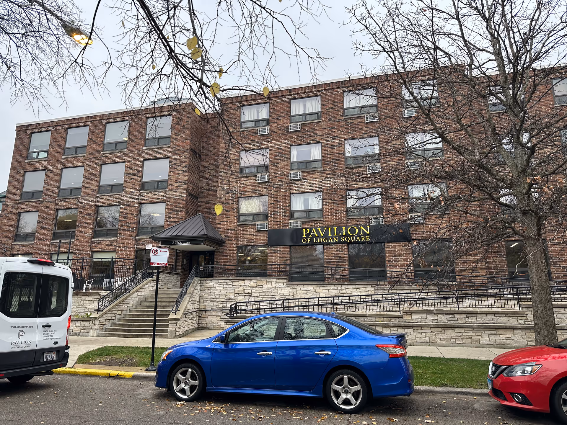 Front view of a brick multi-story building labeled "Pavilion of Logan Square" with cars parked along the street.
