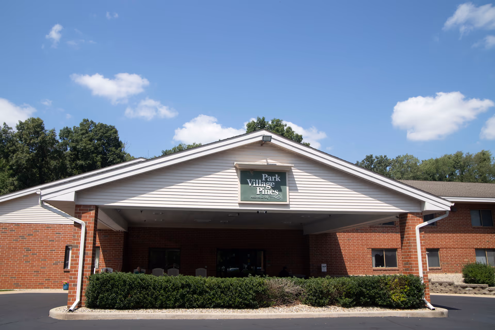 Front exterior view of the Park Village Pines facility showing a brick building with a covered entrance, green bushes in front, and a clear blue sky with some clouds.