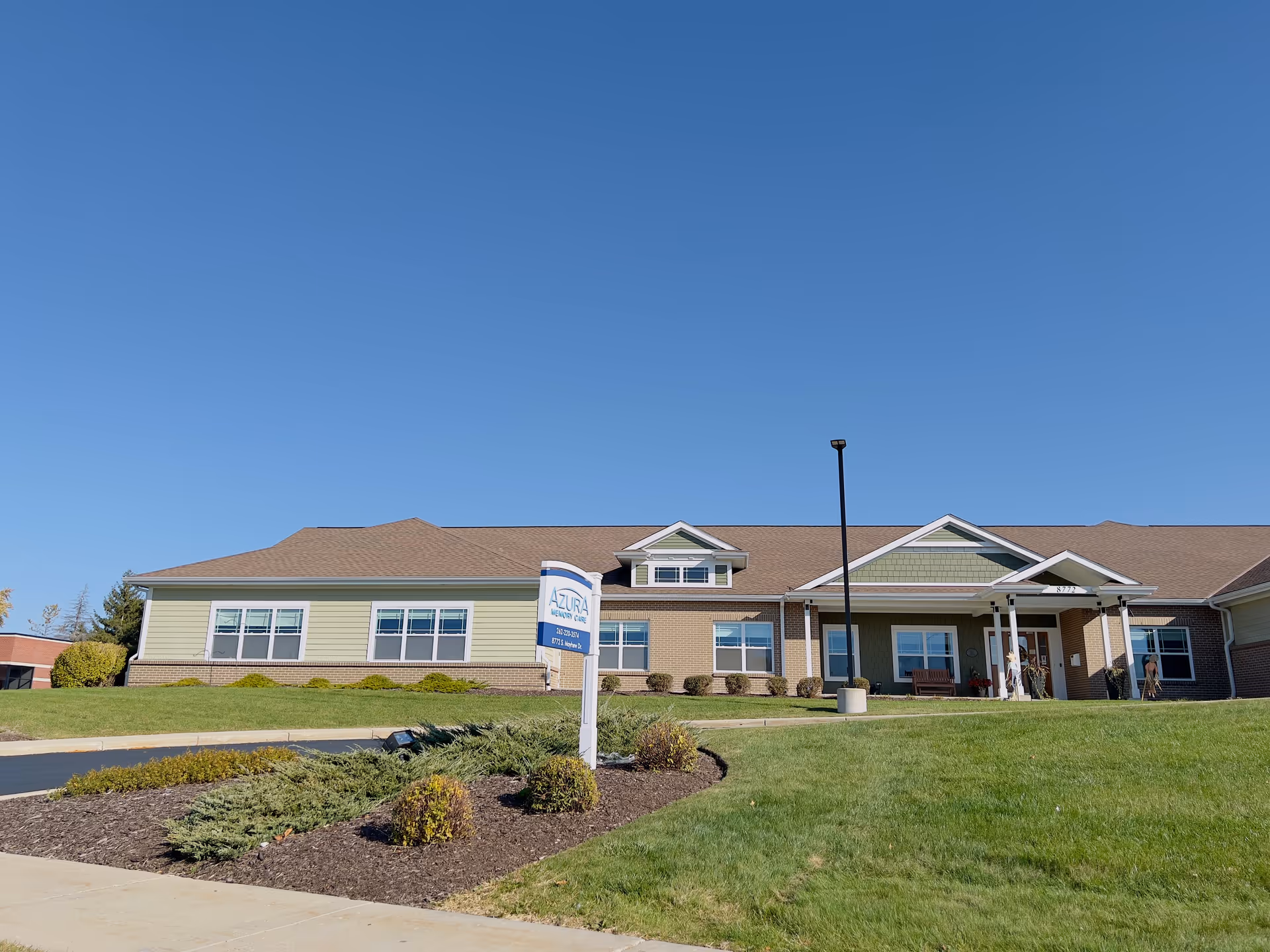 Exterior view of Azura Memory Care of Oak Creek building on a clear sunny day with a well-maintained lawn and landscaping in front. The building has a combination of green siding and brick with multiple windows and a covered entrance.