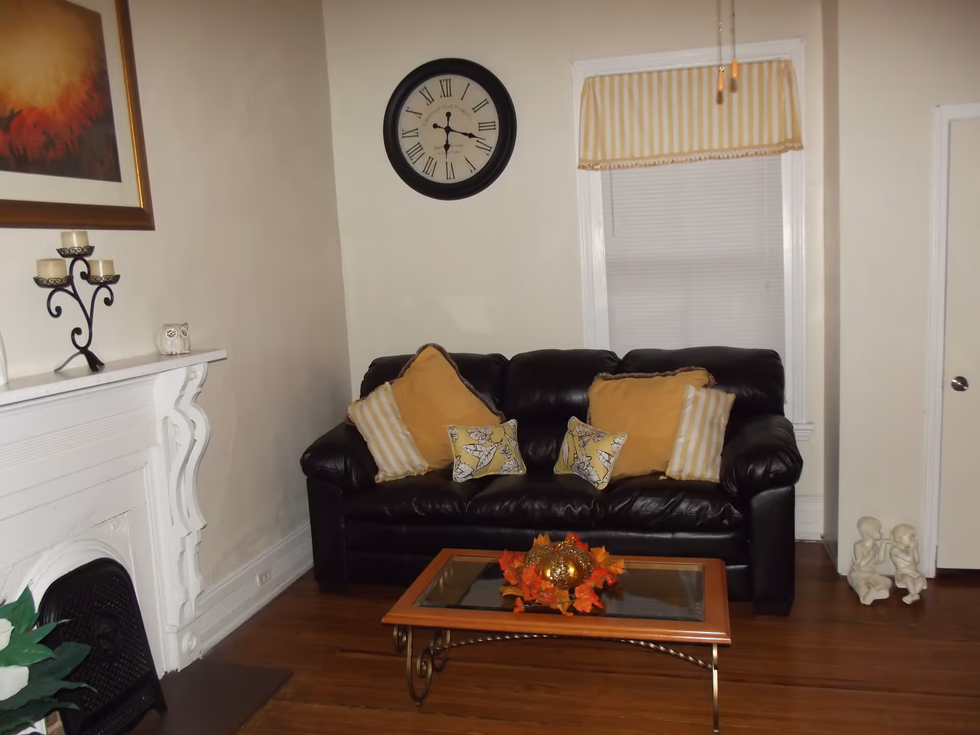 A cozy living room with a black leather couch adorned with yellow and striped pillows. A glass coffee table with a decorative centerpiece sits in front of the couch. On the left, there is a white fireplace mantel with candles and a small owl figurine. A large round clock hangs on the wall above the couch, and a window with a yellow and white striped valance is to the right. Two small statues of children are placed on the floor near a closed white door.