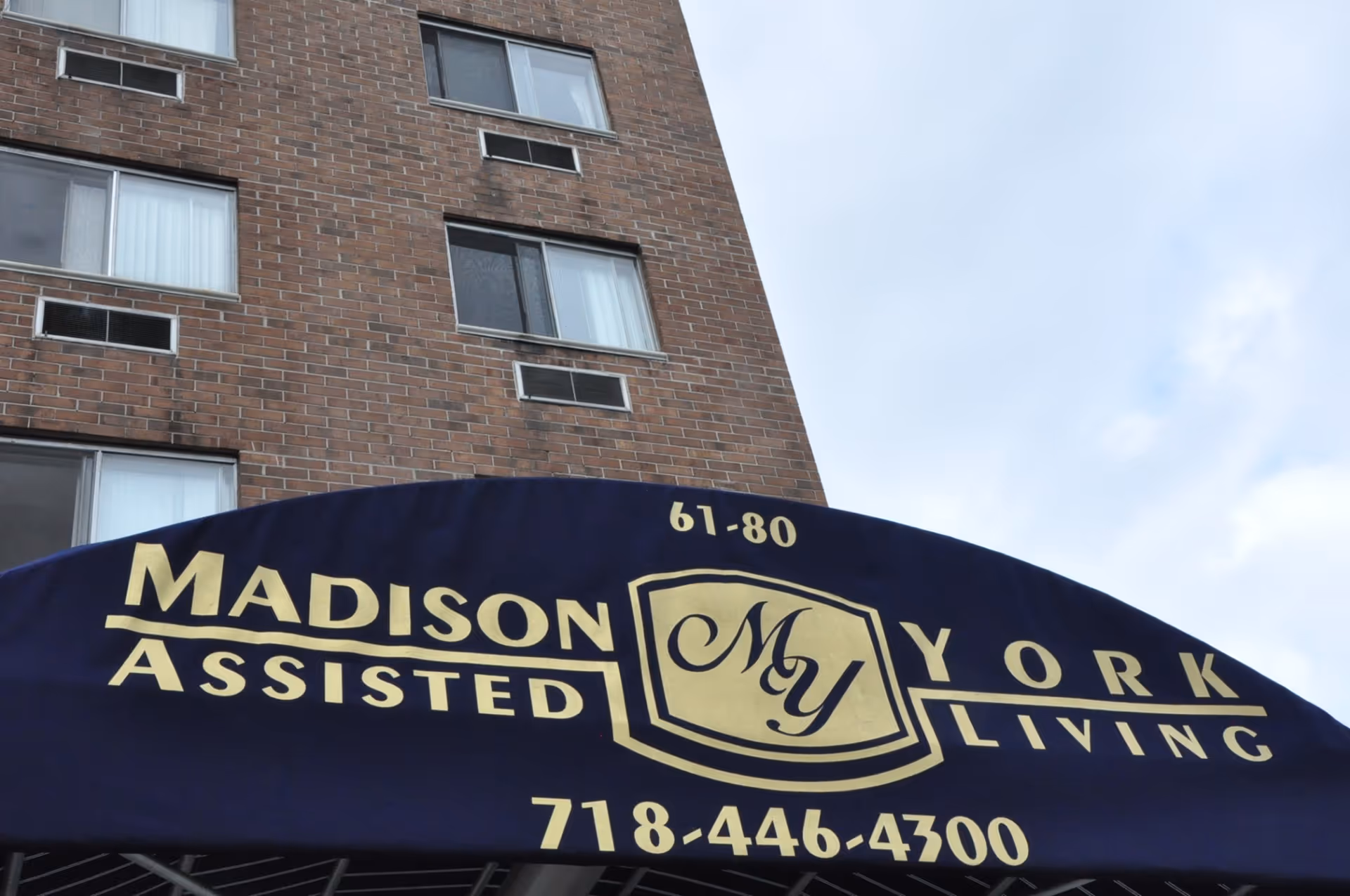 Blue awning with gold lettering displaying the name Madison York Assisted Living, along with the address number 61-80 and phone number 718-446-4300, in front of a brick building with several windows.