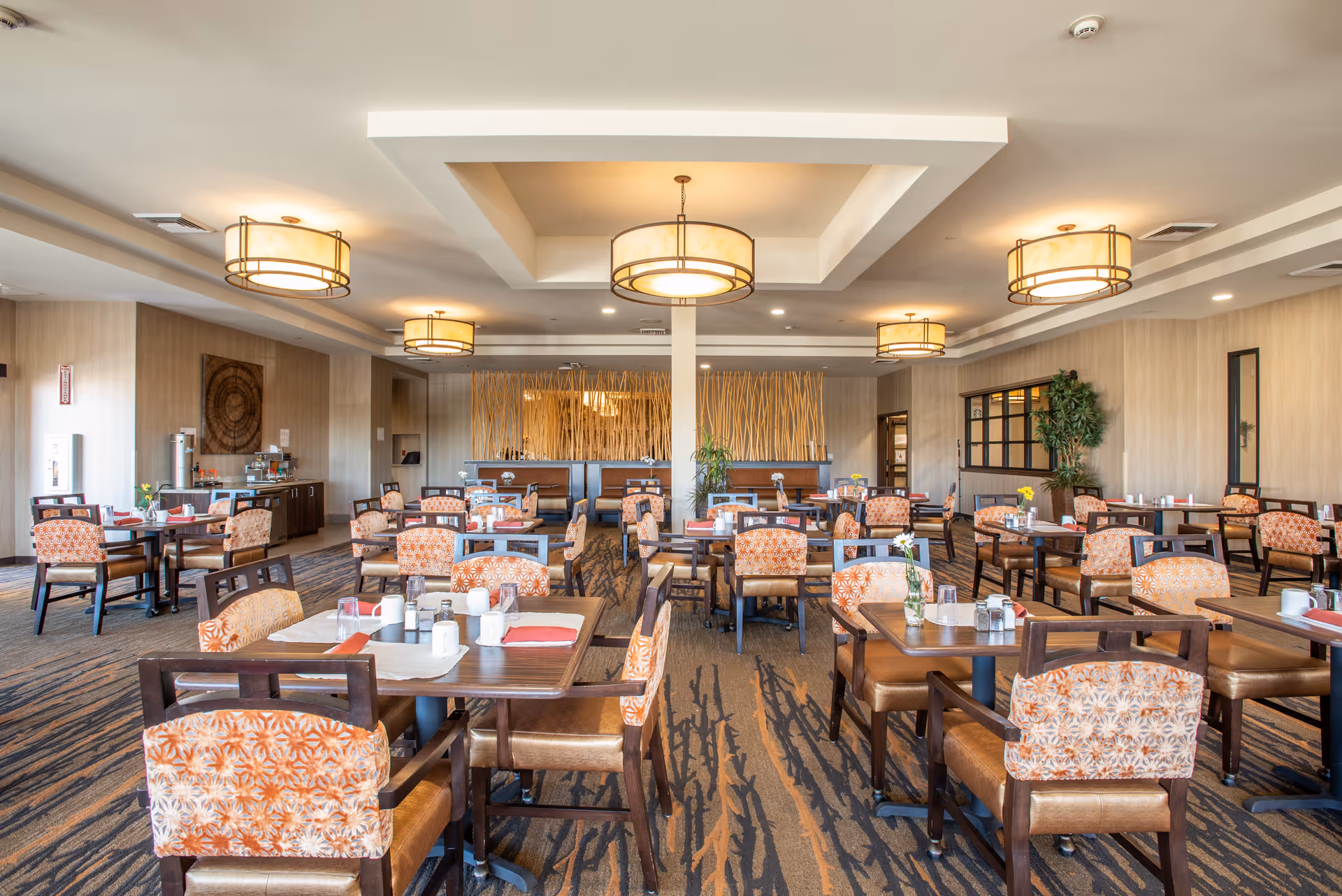 A spacious and well-lit dining room with multiple wooden tables and chairs featuring orange patterned upholstery. The tables are set with placemats, napkins, cups, and small flower vases. The room has modern ceiling lights, a patterned carpet, and decorative elements including a wooden art piece on the wall and tall plants.