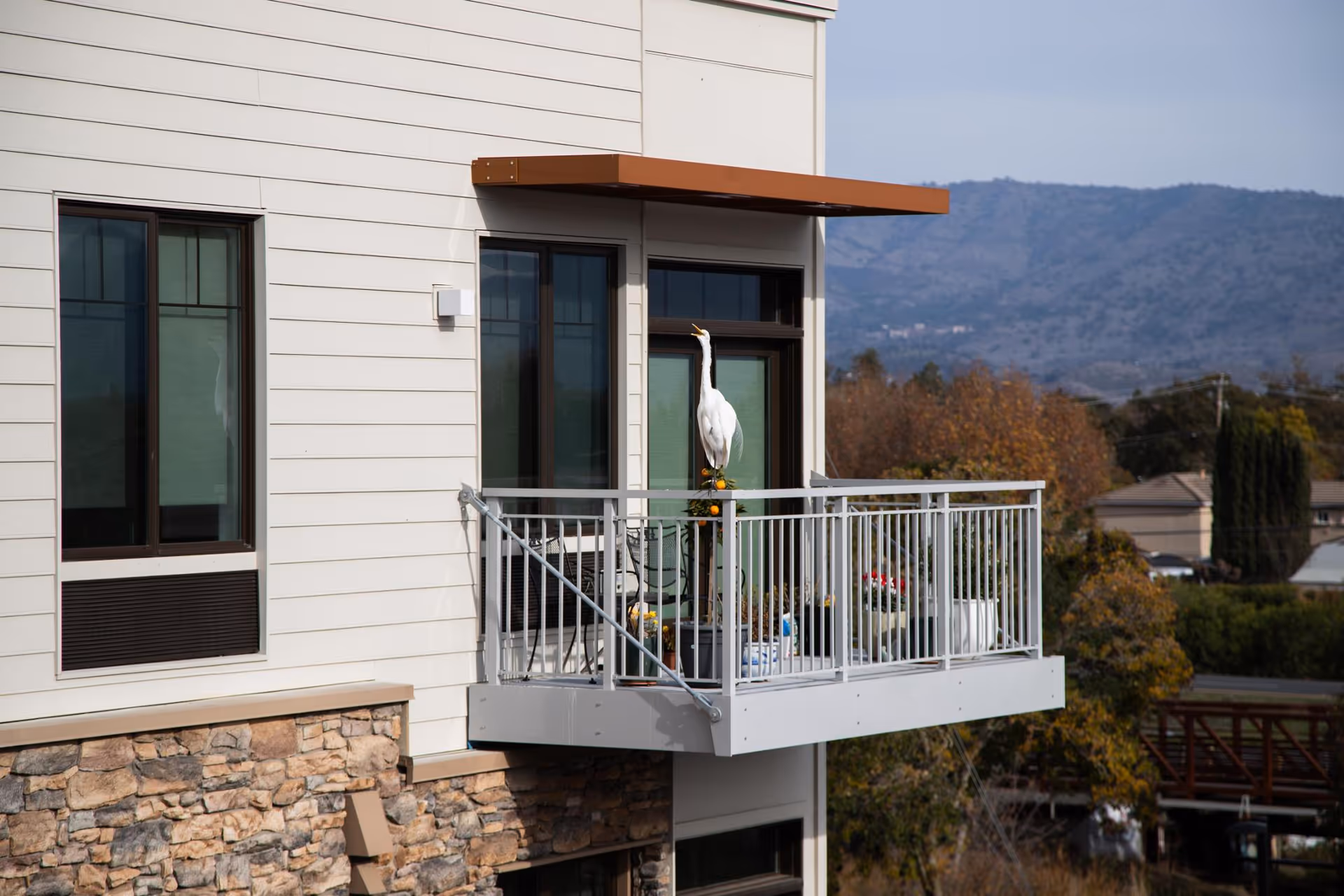 A small balcony attached to a building with white siding and stone accents. The balcony has a metal railing and contains a large white bird statue, some potted plants, and outdoor furniture. In the background, there are trees, houses, and hills under a clear sky.