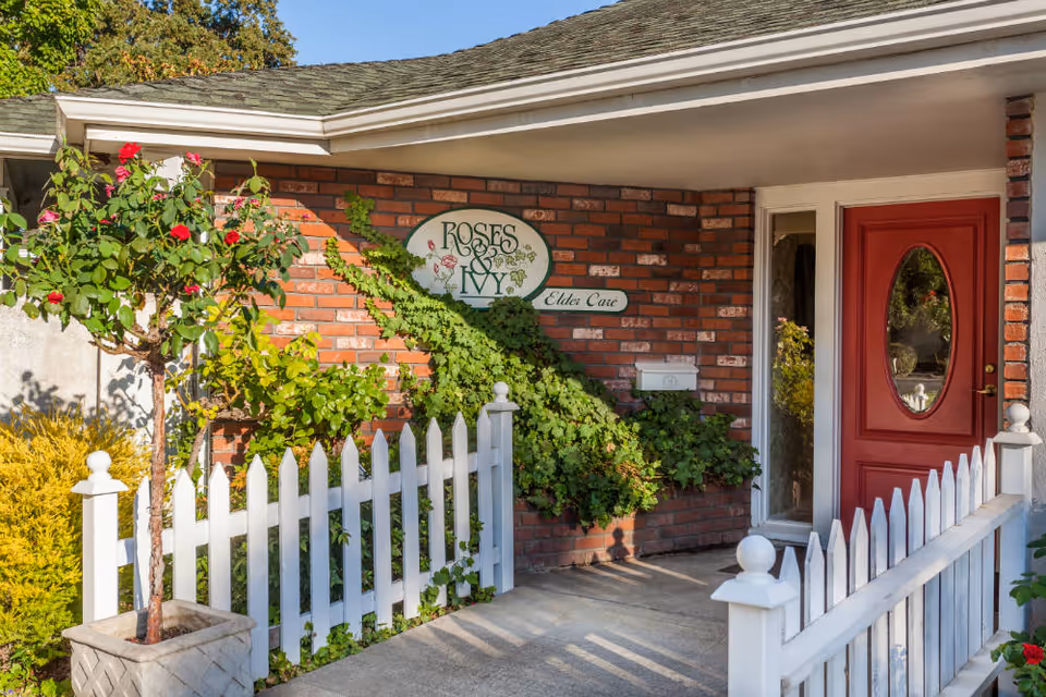 Entrance of Roses & Ivy Assisted Living Facility for the Elderly showing a red door with an oval window, a white picket fence, a potted rose plant, and a brick wall with a sign that reads 'Roses & Ivy Elder Care' partially covered by ivy.