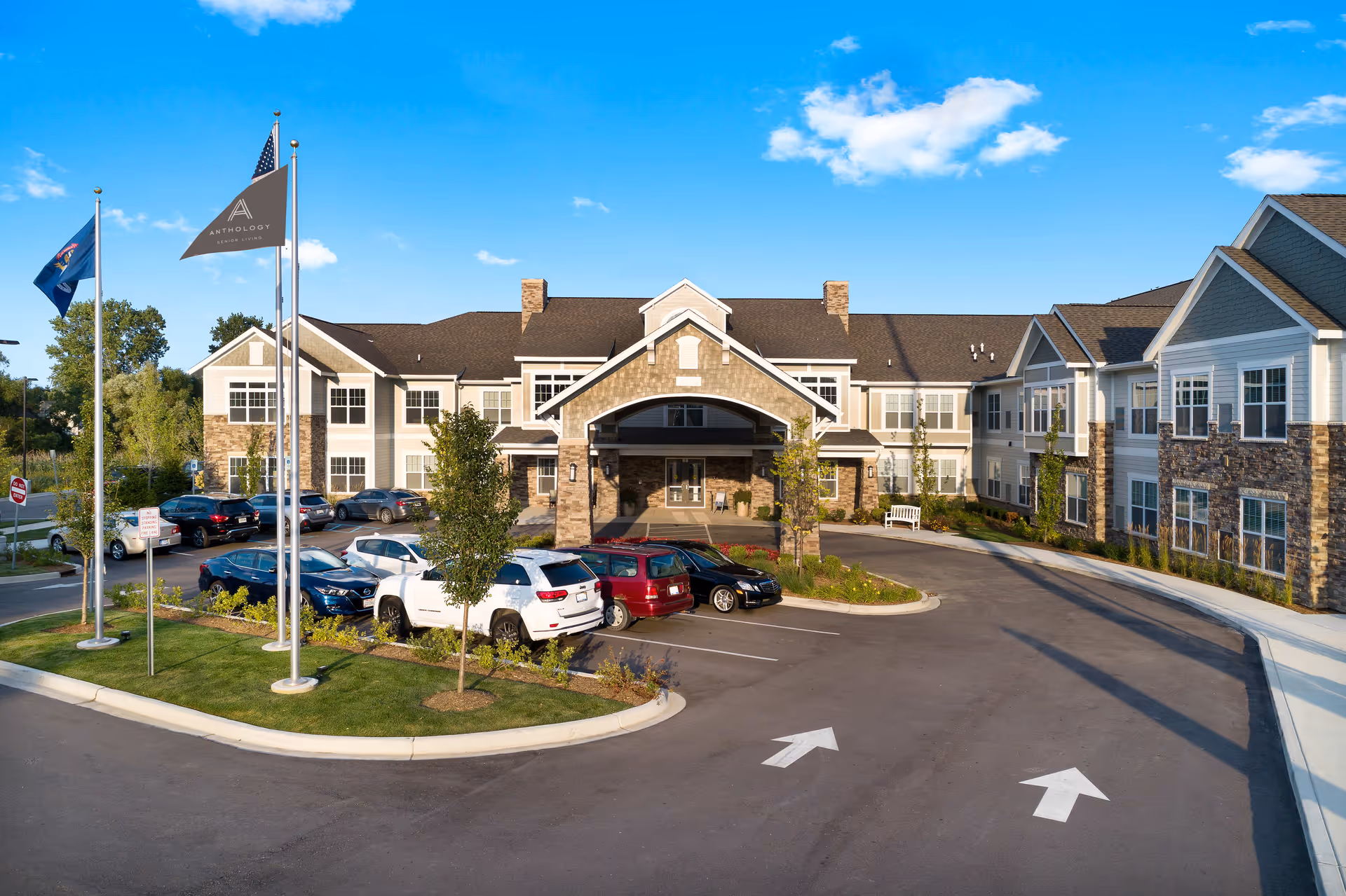 Exterior view of a senior living facility building with a driveway and parking lot in front. The building has a stone and siding facade with multiple windows and a covered entrance. Three flagpoles with flags are visible on the left side of the image. Several cars are parked in the lot under a clear blue sky.