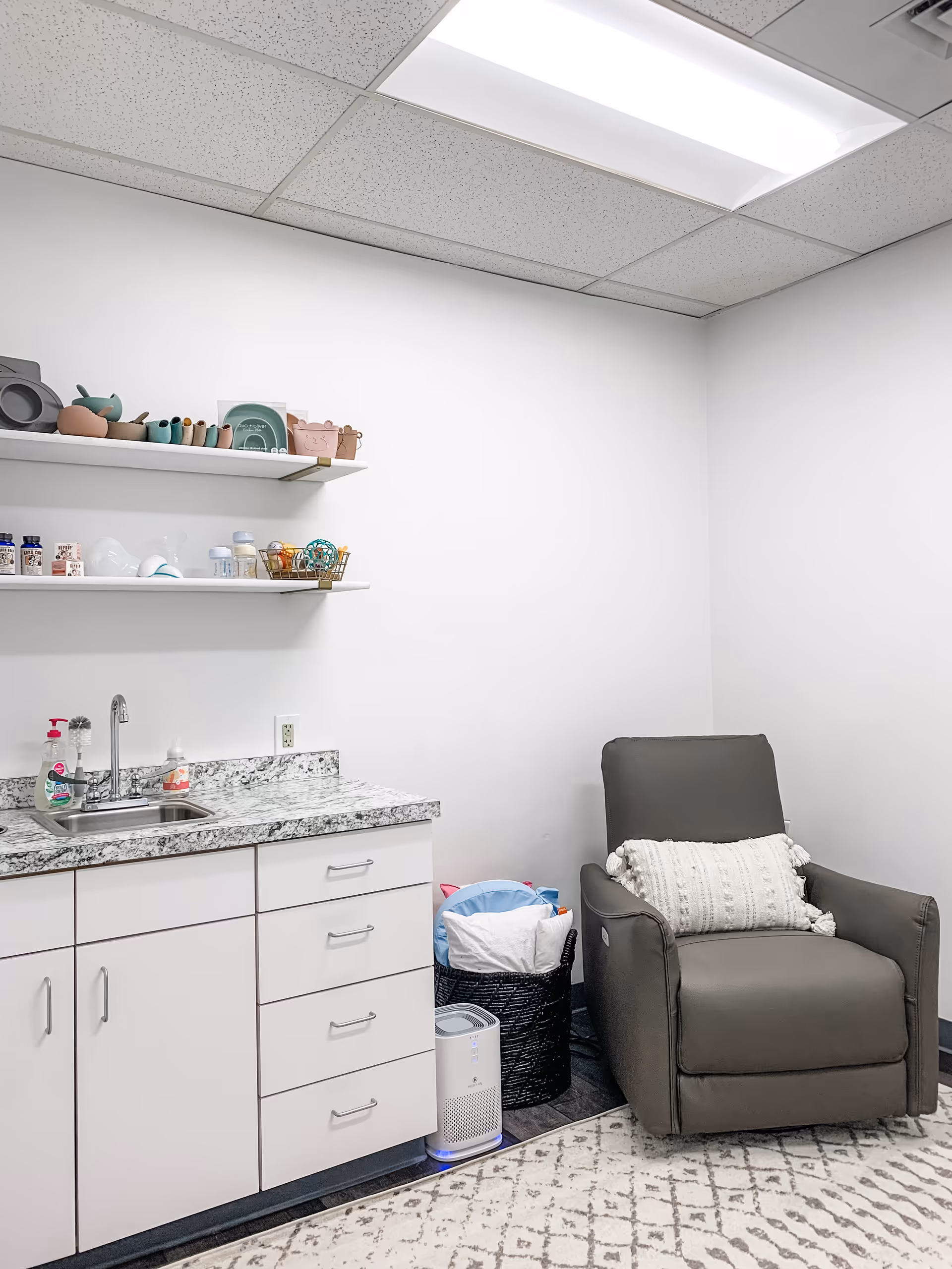 Small, clean kitchenette with sink, white cabinets, shelves of dishes, and a gray armchair.