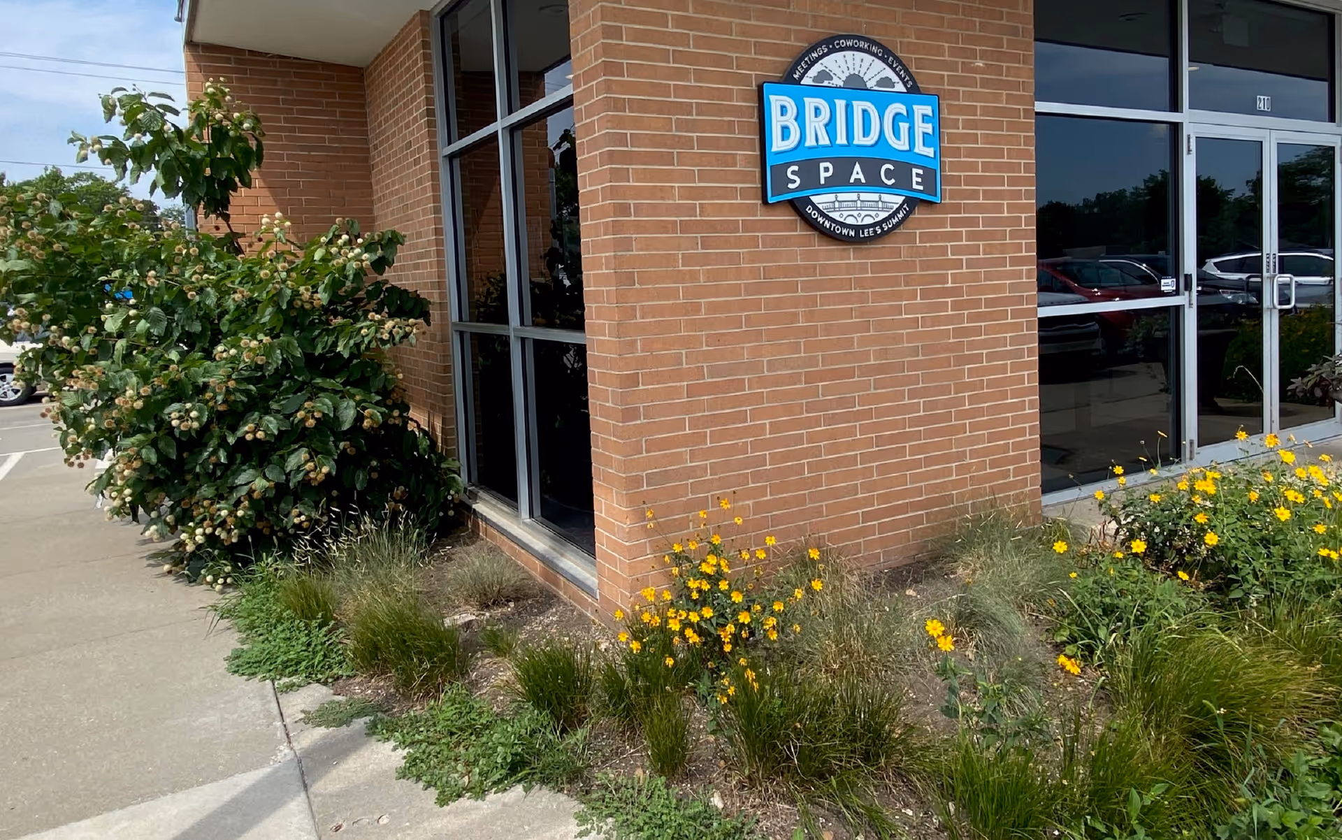 Exterior brick building with Bridge Space sign and glass doors, surrounded by plants.