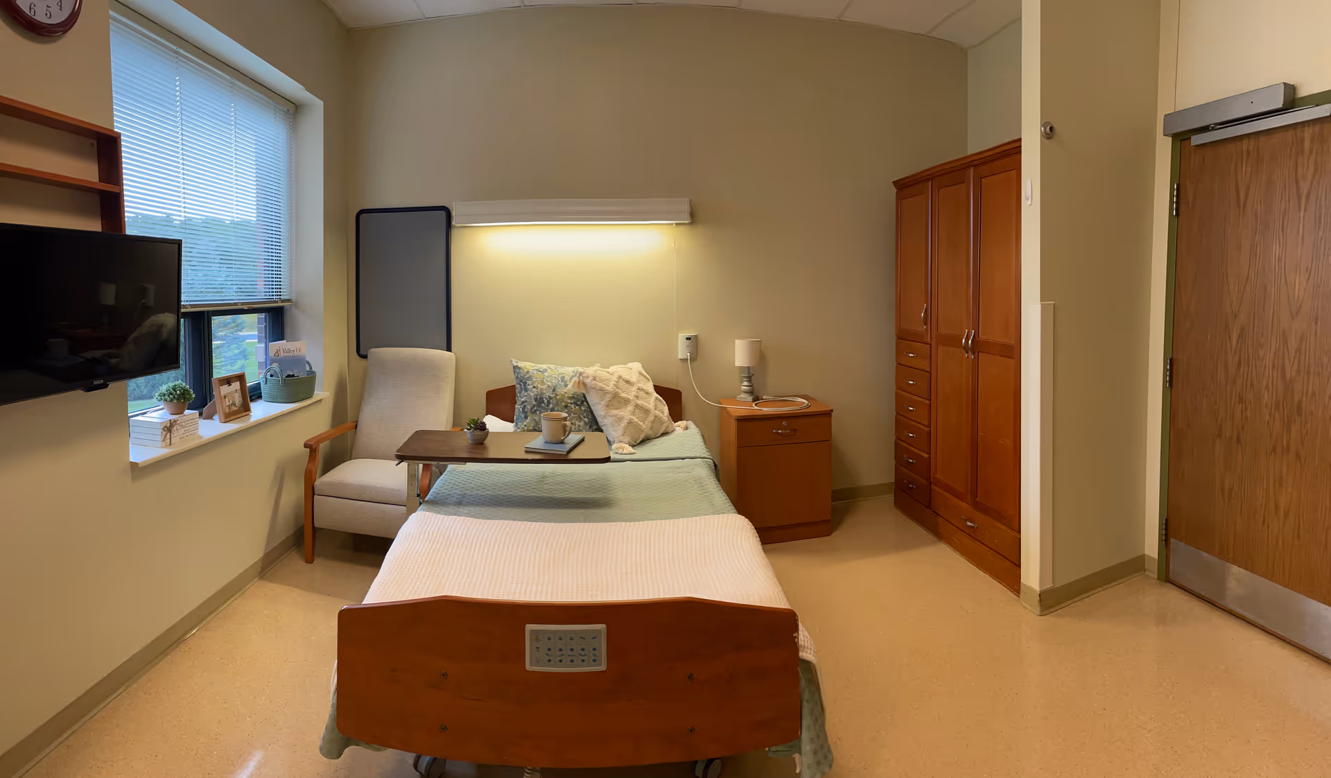 A nursing facility bedroom with a single bed made with pillows and blankets, a wooden nightstand with a lamp, a wooden wardrobe, a comfortable armchair, a small table with a cup and book, a wall-mounted TV, and a window with blinds letting in natural light.