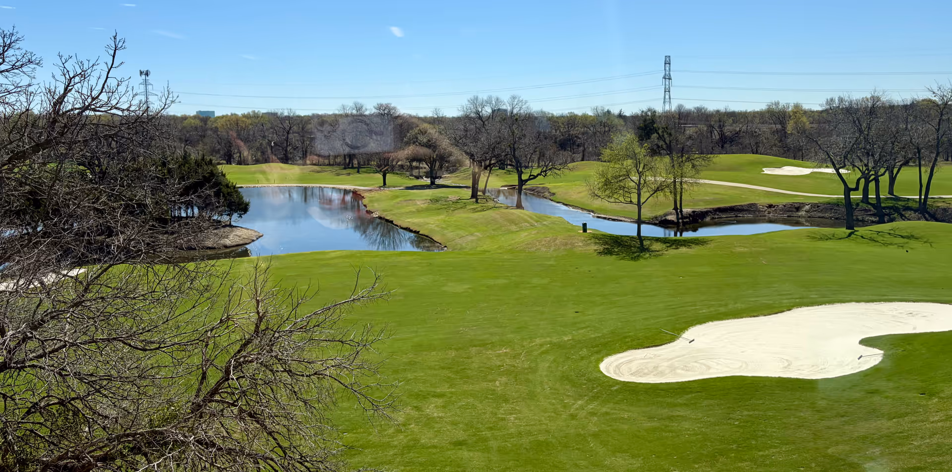 Sunlit golf course with green fairways, trees, water hazards, and a sandy bunker.