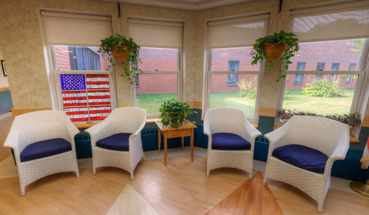 Sunlit seating area with four white wicker chairs with blue cushions, potted plants, and large windows overlooking a brick courtyard.