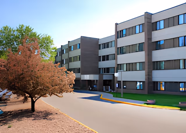 Exterior view of a multi-story senior living facility building with several windows, a tree with reddish leaves on the left side, and a paved driveway in front.