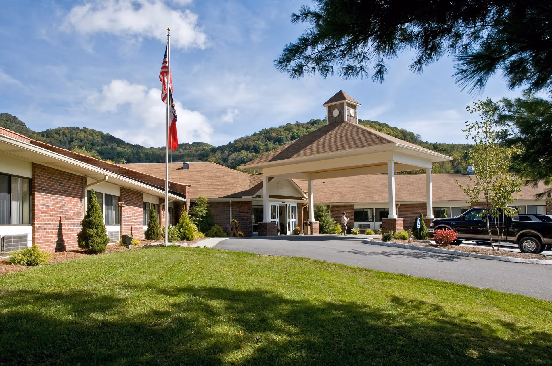Single-story brick building with a covered entrance/porte-cochere, flagpole, parked vehicle and tree-covered hills in the background.