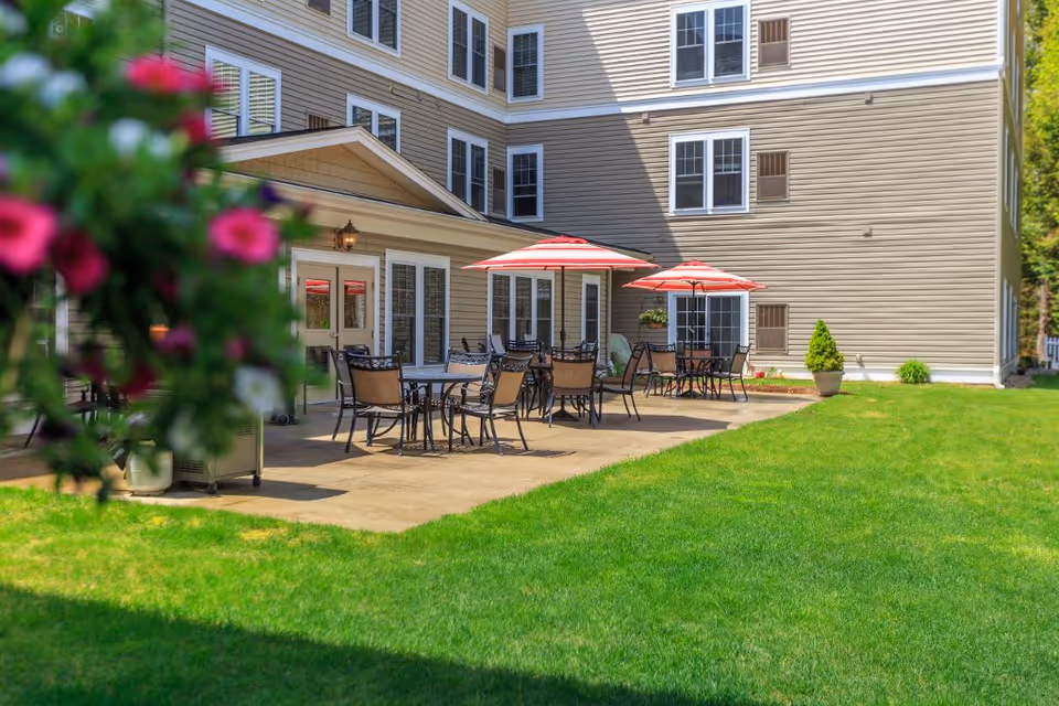 Outdoor patio area at Bellamy Fields Assisted Living with several tables and chairs under red and white striped umbrellas, adjacent to a beige multi-story building with multiple windows. Green grass lawn surrounds the patio, and some pink flowers are visible in the foreground.