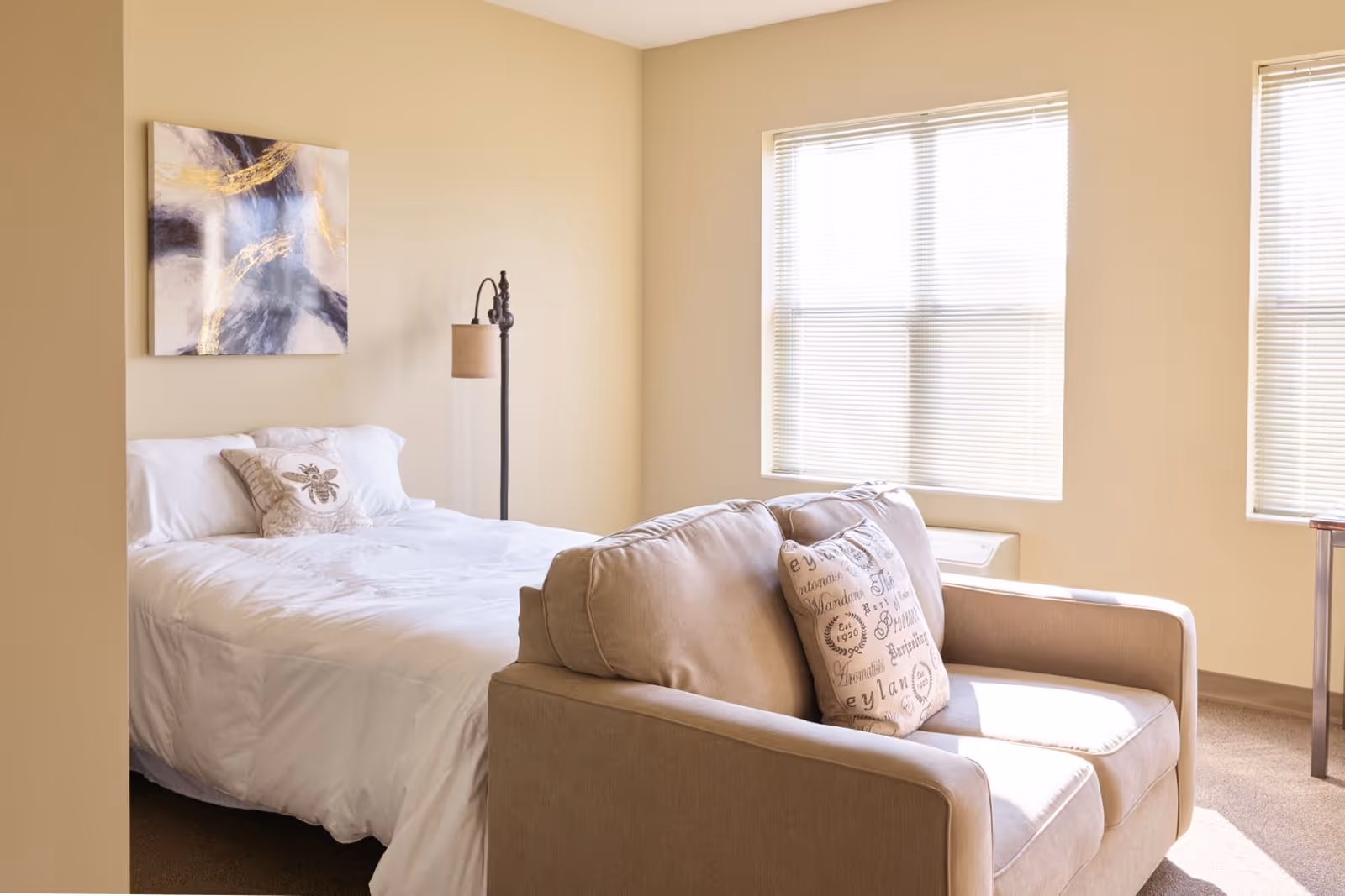 A bright bedroom with a neatly made bed featuring white bedding and decorative pillows. A beige loveseat with patterned pillows is positioned at the foot of the bed. The room has light-colored walls, two windows with blinds allowing natural light to enter, a floor lamp beside the bed, and a piece of abstract artwork hanging on the wall above the bed.