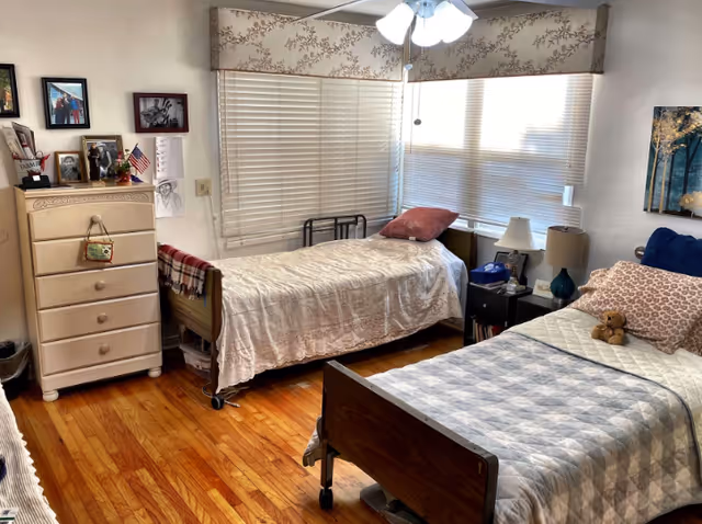 Two single beds in a furnished senior living bedroom with dresser, photos, and windows.