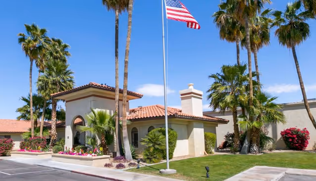 Front exterior of a single-story stucco building with a red tile roof, palm trees, landscaped grounds and an American flag.