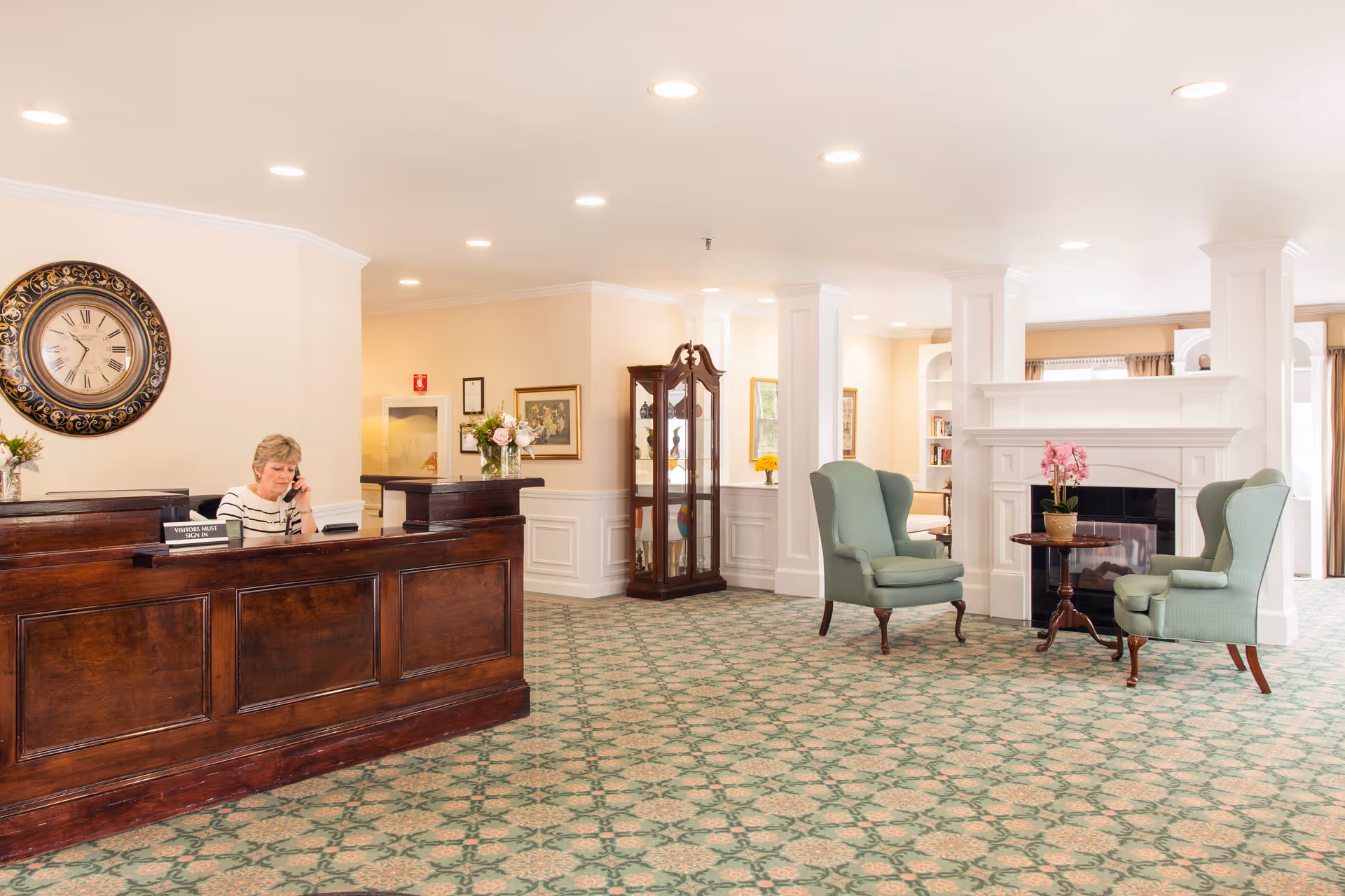 Reception area of Royal Health Braintree featuring a woman sitting behind a dark wooden reception desk talking on the phone. The room has a large decorative wall clock, floral arrangements, a patterned carpet, two green upholstered armchairs with a small round table and a potted orchid between them, and a white fireplace with built-in shelves.