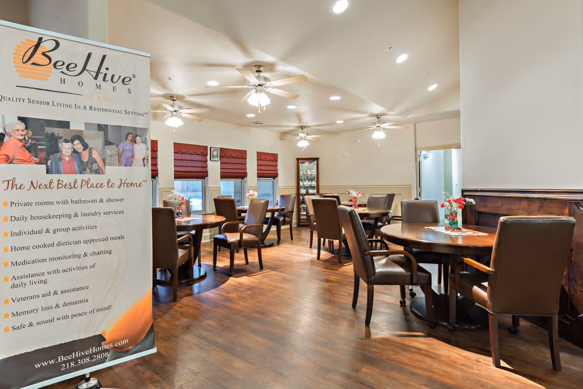 Dining room in a BeeHive senior living facility with round tables, chairs, ceiling fans, and a promotional banner at left.