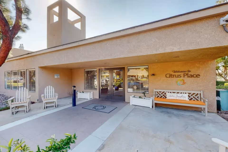 Exterior entrance of Citrus Place facility with a covered walkway, two white wooden chairs on the left, a bench with an orange cushion and a 'Hello Sunshine' pillow on the right, and glass double doors in the center. The building has a beige stucco finish and a small tower structure above the roof.