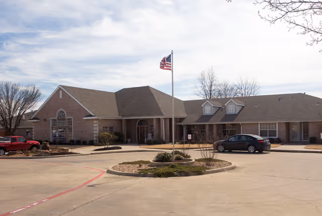 Exterior view of a single-story brick building with a peaked roof, an American flag on a flagpole in front, a circular driveway, and parked cars under a partly cloudy sky.
