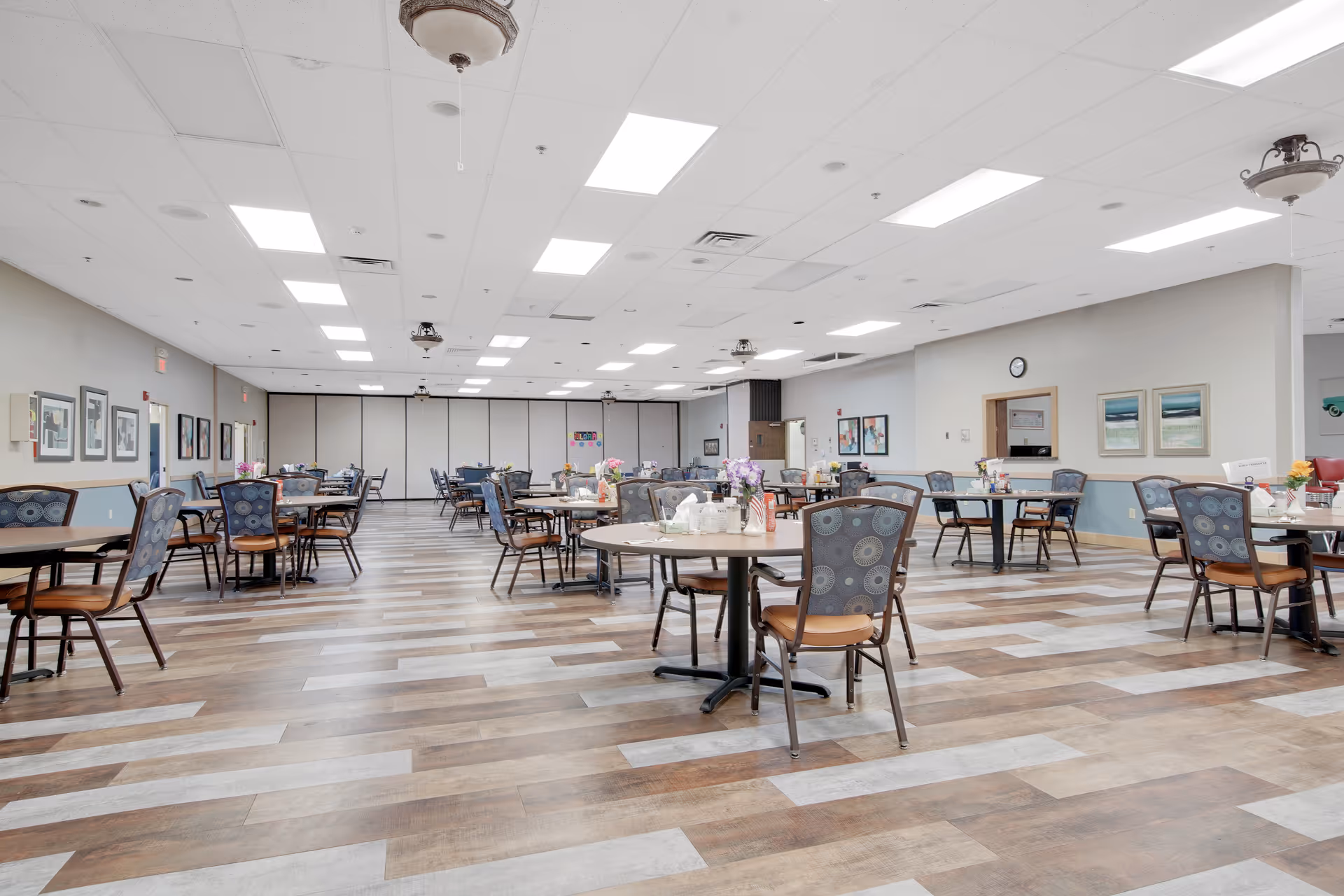A spacious dining room with multiple round tables and chairs arranged neatly. The room has a light-colored ceiling with recessed lighting, wood-patterned flooring, and framed artwork on the walls. Some tables have flower centerpieces and condiments.