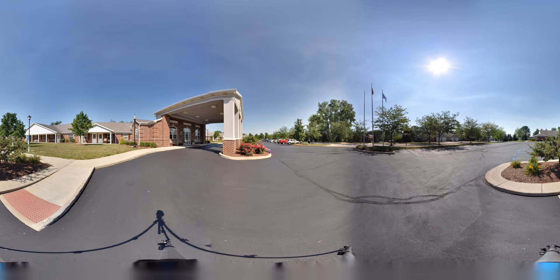 Exterior view of Five Star Residences of Fort Wayne showing the main entrance with a covered drop-off area, a paved driveway, landscaped areas with trees and bushes, and a parking lot under a clear blue sky with the sun shining.