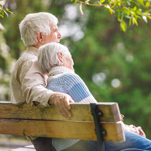 Two elderly people sit on a wooden outdoor bench, enjoying a serene park.