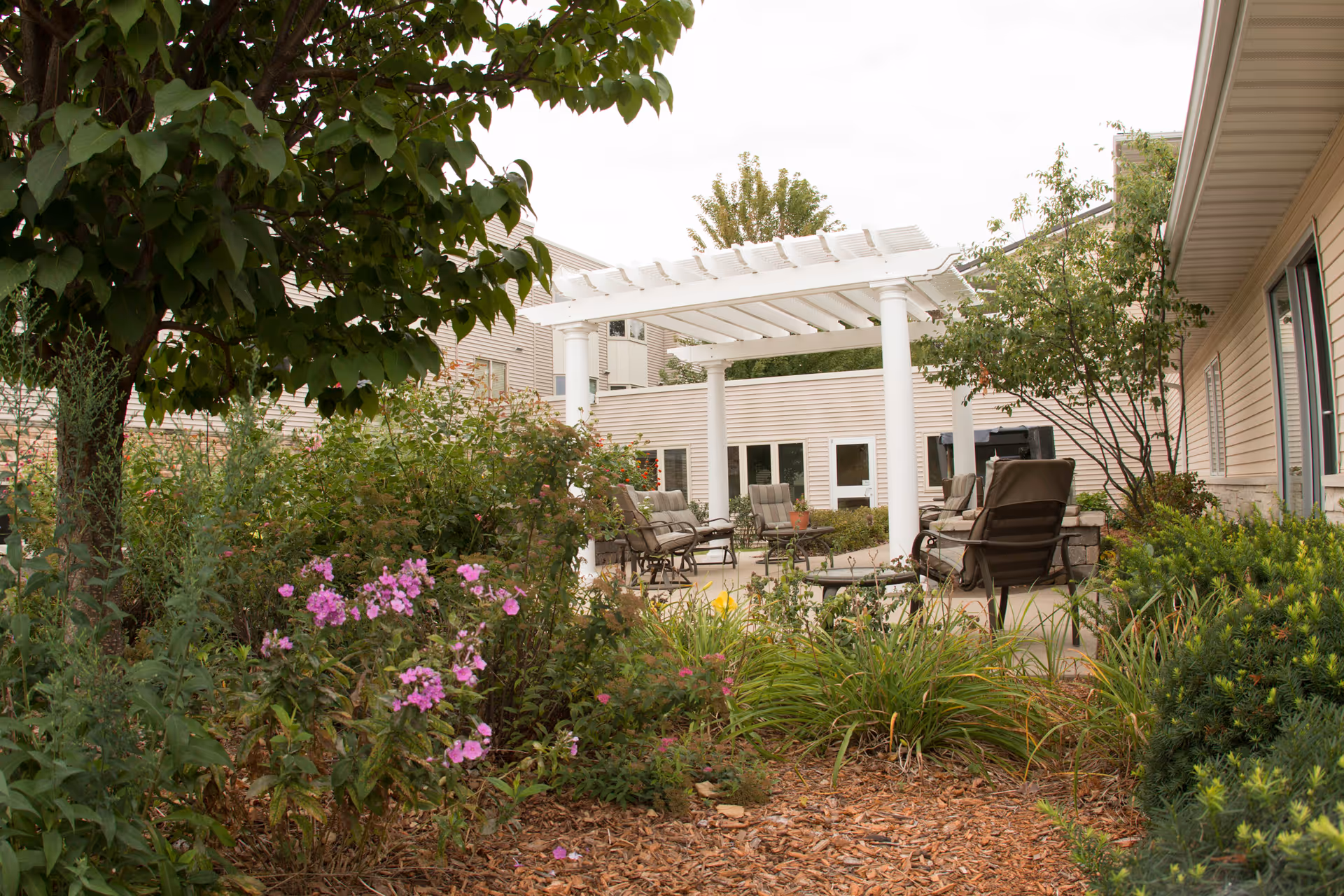 Outdoor patio area at The Views of Cedar Rapids | RidgeView Assisted Living featuring a white pergola, several cushioned chairs and tables, surrounded by green shrubs, flowering plants, and trees.