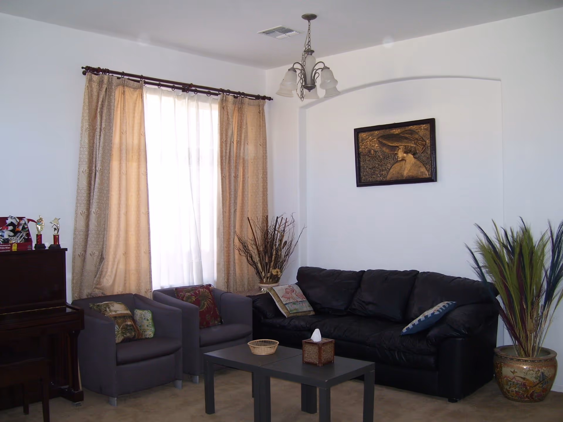 A living room with a black leather couch and two gray armchairs with decorative pillows. A small coffee table with a tissue box and a basket is in front of the seating. Beige curtains cover a window behind the armchairs. A piano with trophies on top is on the left side. A framed artwork of a woman wearing a large hat hangs on the white wall above the couch. A decorative plant in a pot is on the right side of the couch.