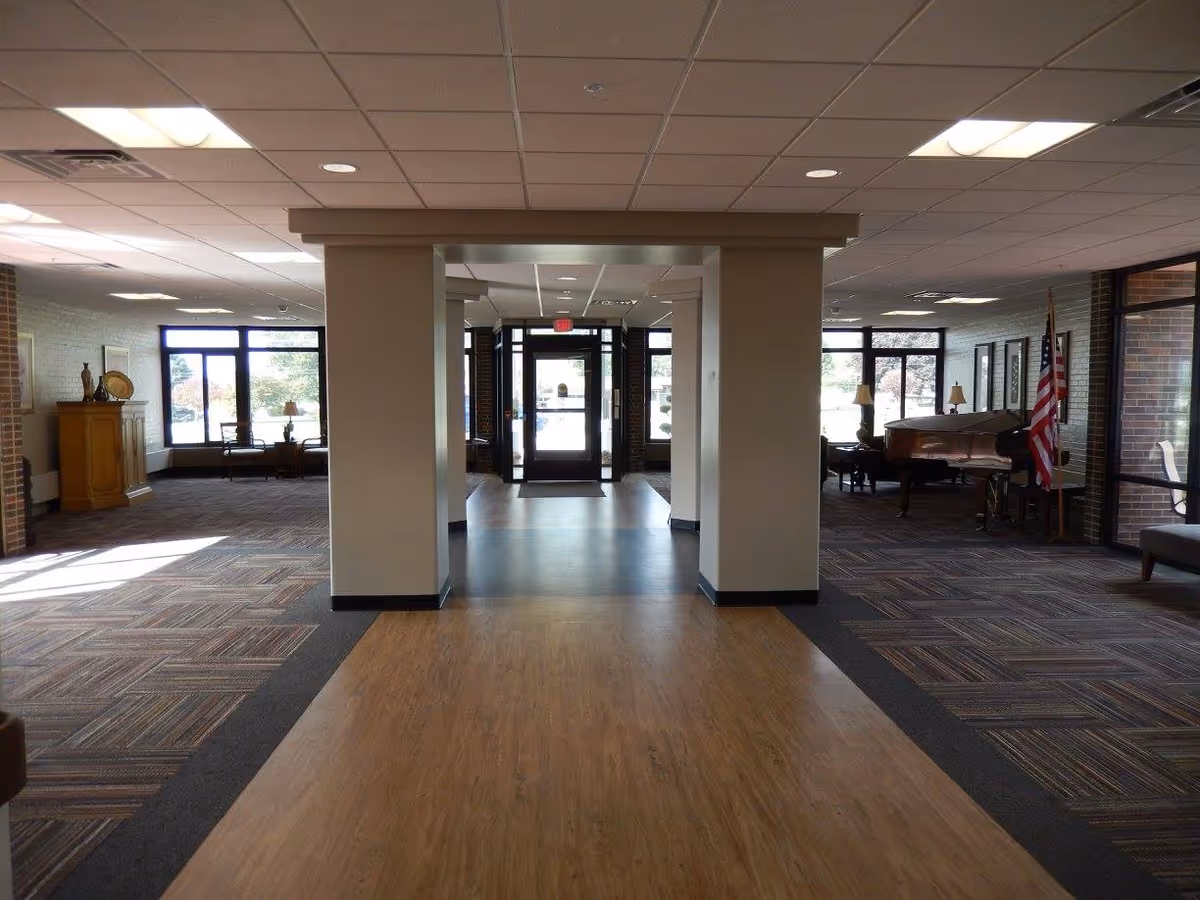 Interior view of a senior living facility lobby with a wooden floor pathway leading to a glass entrance door. On either side of the pathway are carpeted areas with seating arrangements, lamps, a piano, an American flag, and decorative furniture. Large windows allow natural light to fill the space.