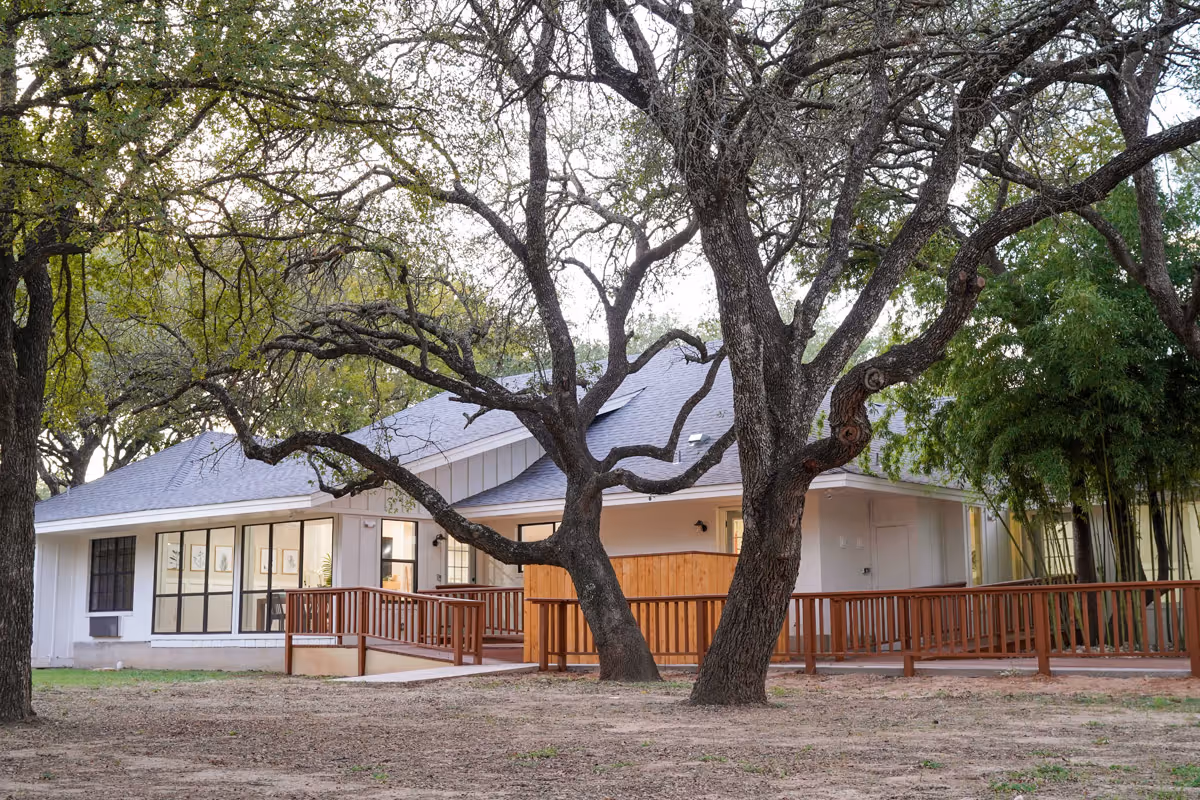 Single-story building exterior with large oak trees and a wooden ramp and porch in front.