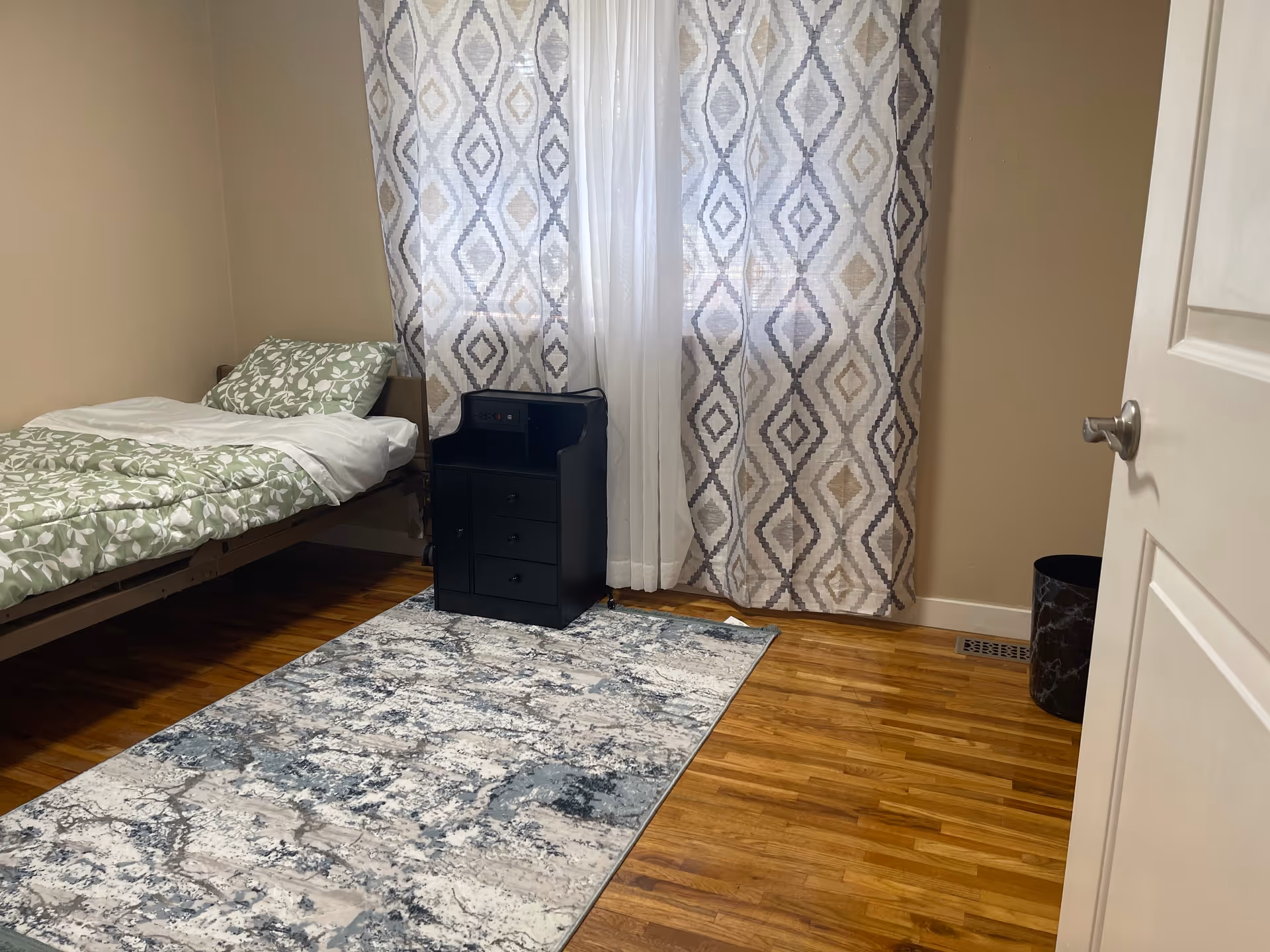A single bedroom features a bed, dresser, patterned curtains, and a blue-gray rug.