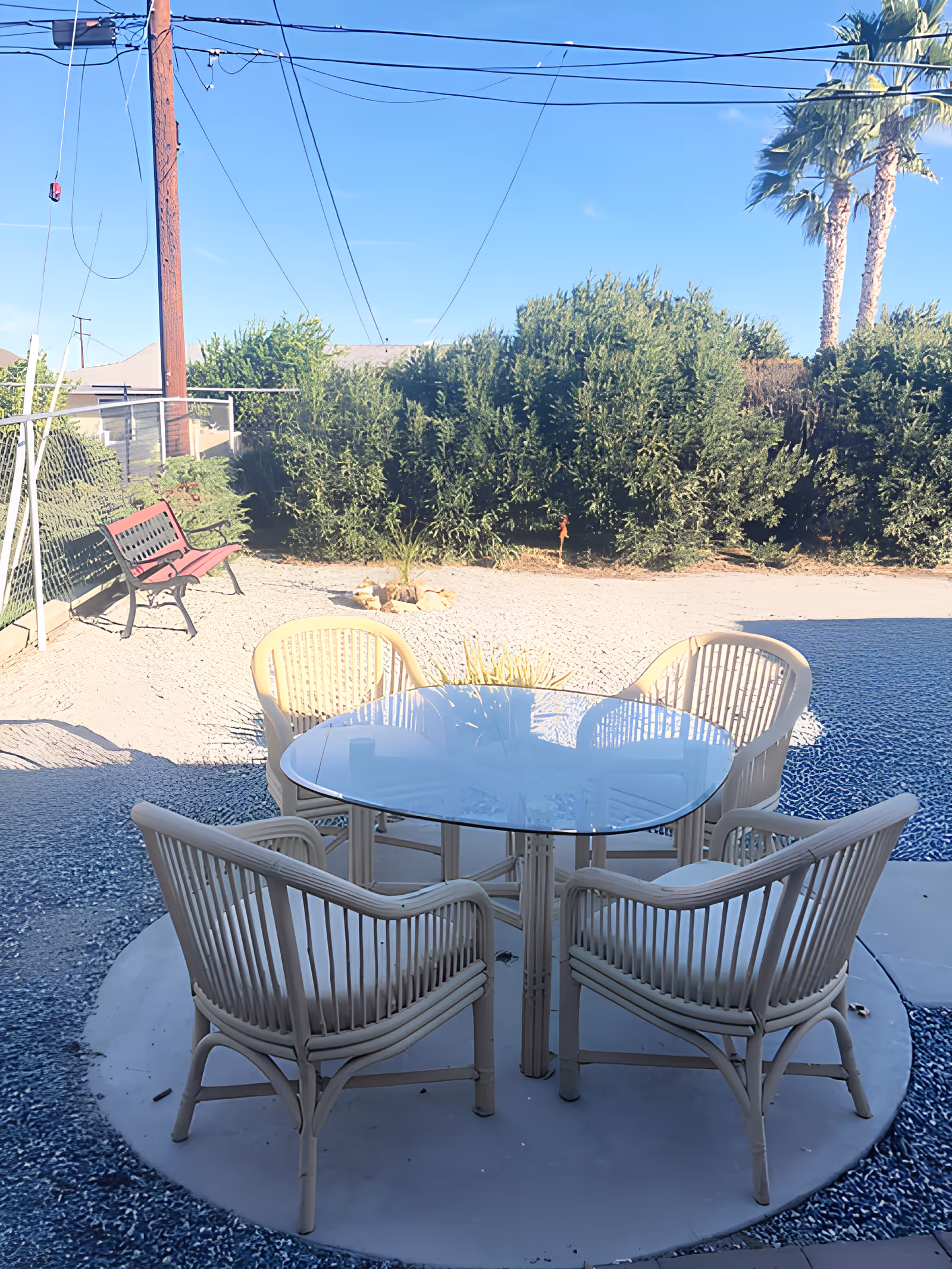 Patio with a round glass-top table and four wicker chairs on a circular pad in a gravel backyard with shrubs and palm trees.