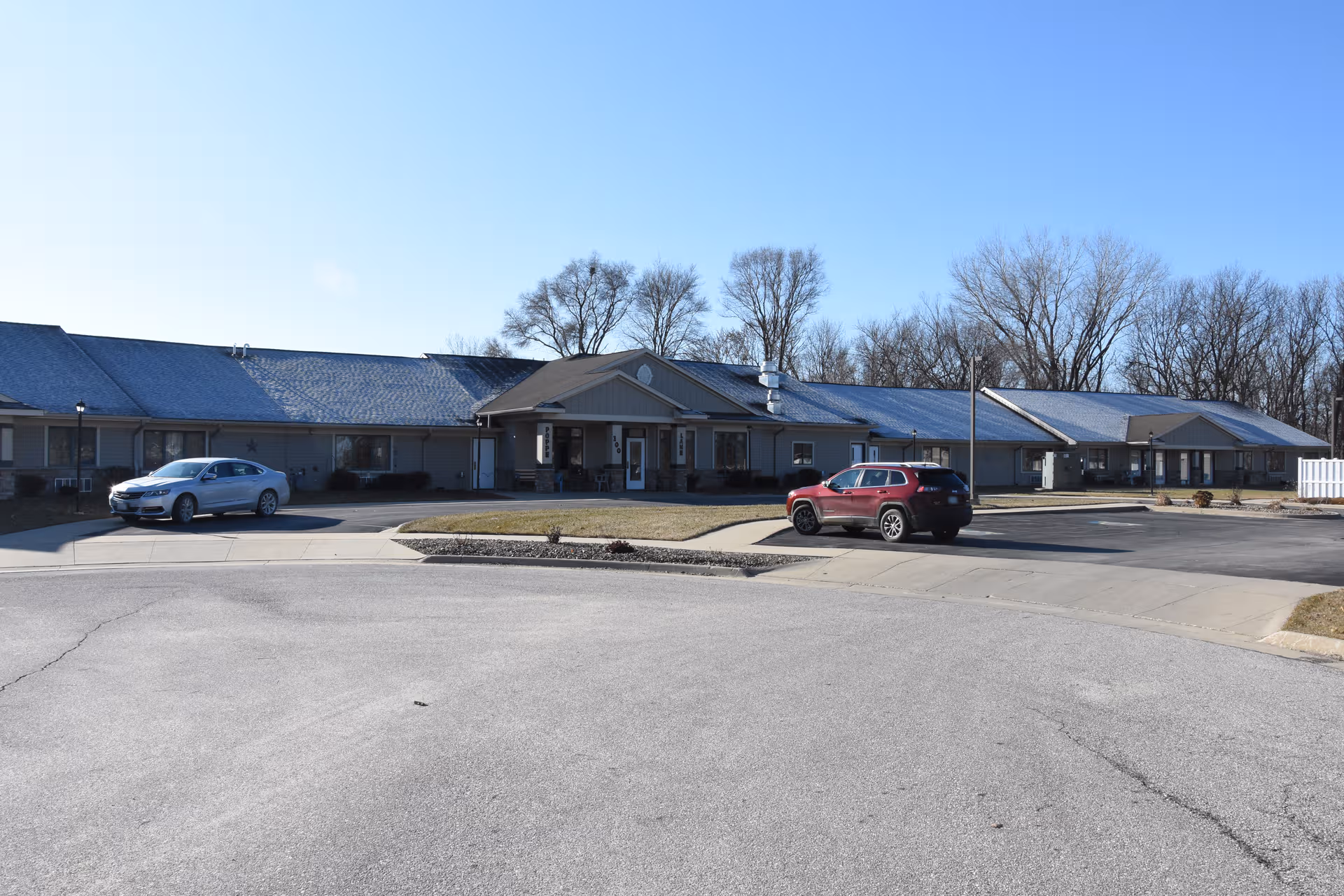 Single-story senior living facility front with parking lot and leafless trees in background.