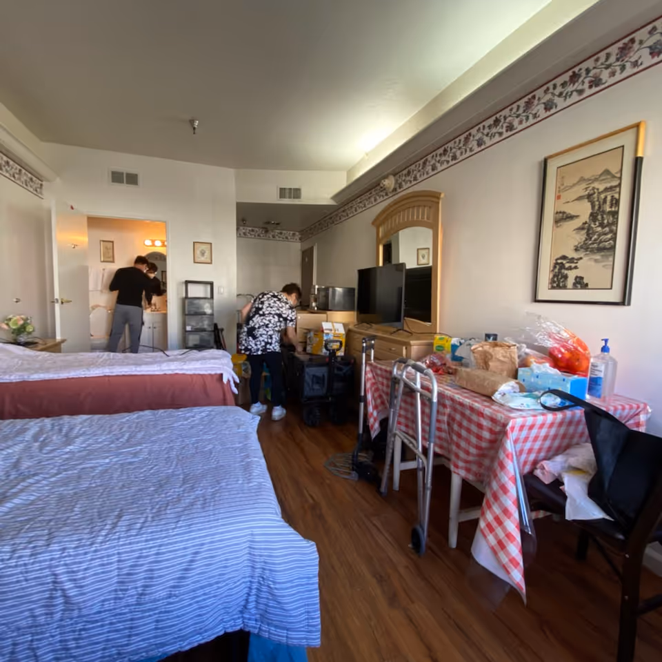 Indoor senior living room with two beds, a checkered dining table, and people nearby.