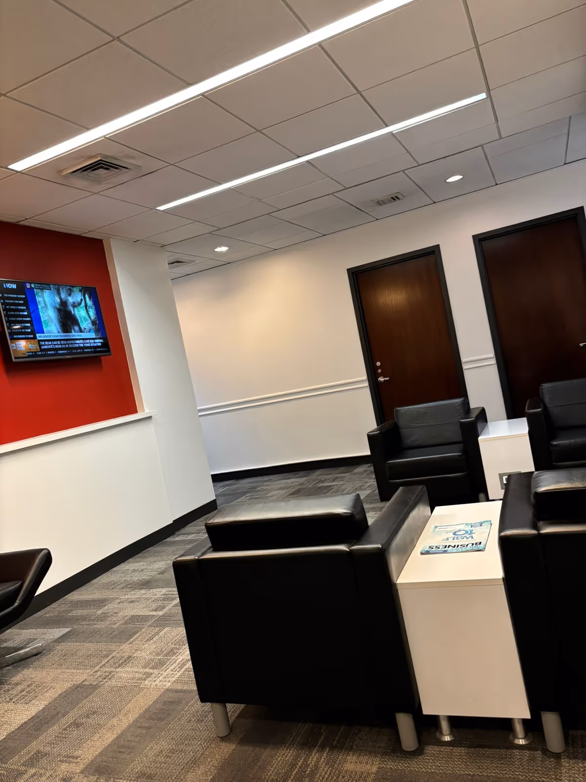 Modern waiting lounge with black leather chairs, white tables, and a wall-mounted TV.