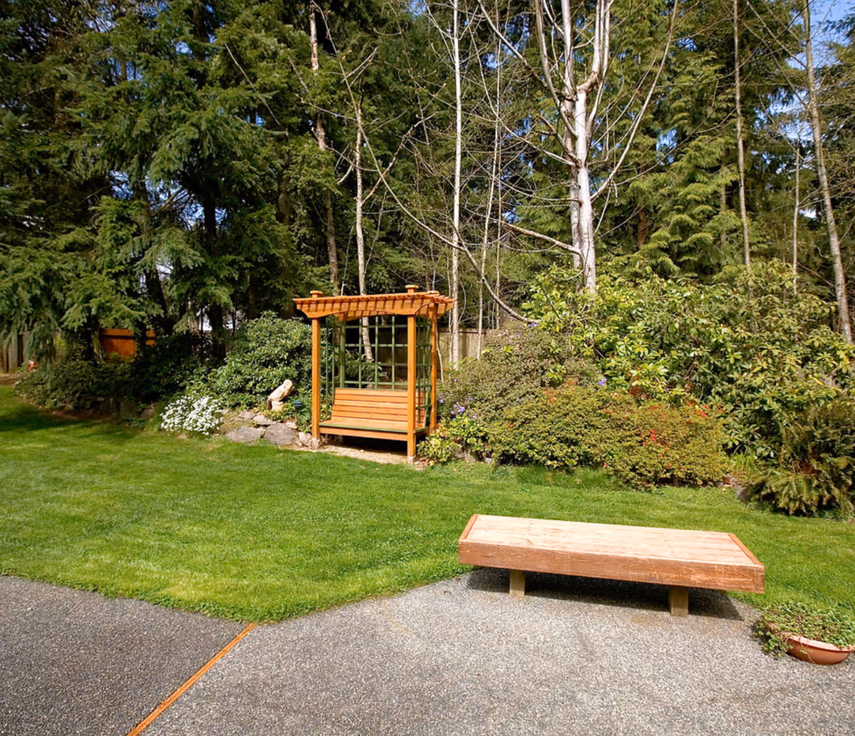 A wooden garden bench beneath a small arbor, surrounded by lush shrubs.
