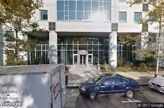 Exterior view of a building entrance with glass doors, columns, and parked cars.