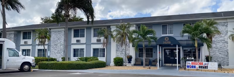 Front exterior of Elite at Cape Coral showing entrance, palm trees, and a parked vehicle
