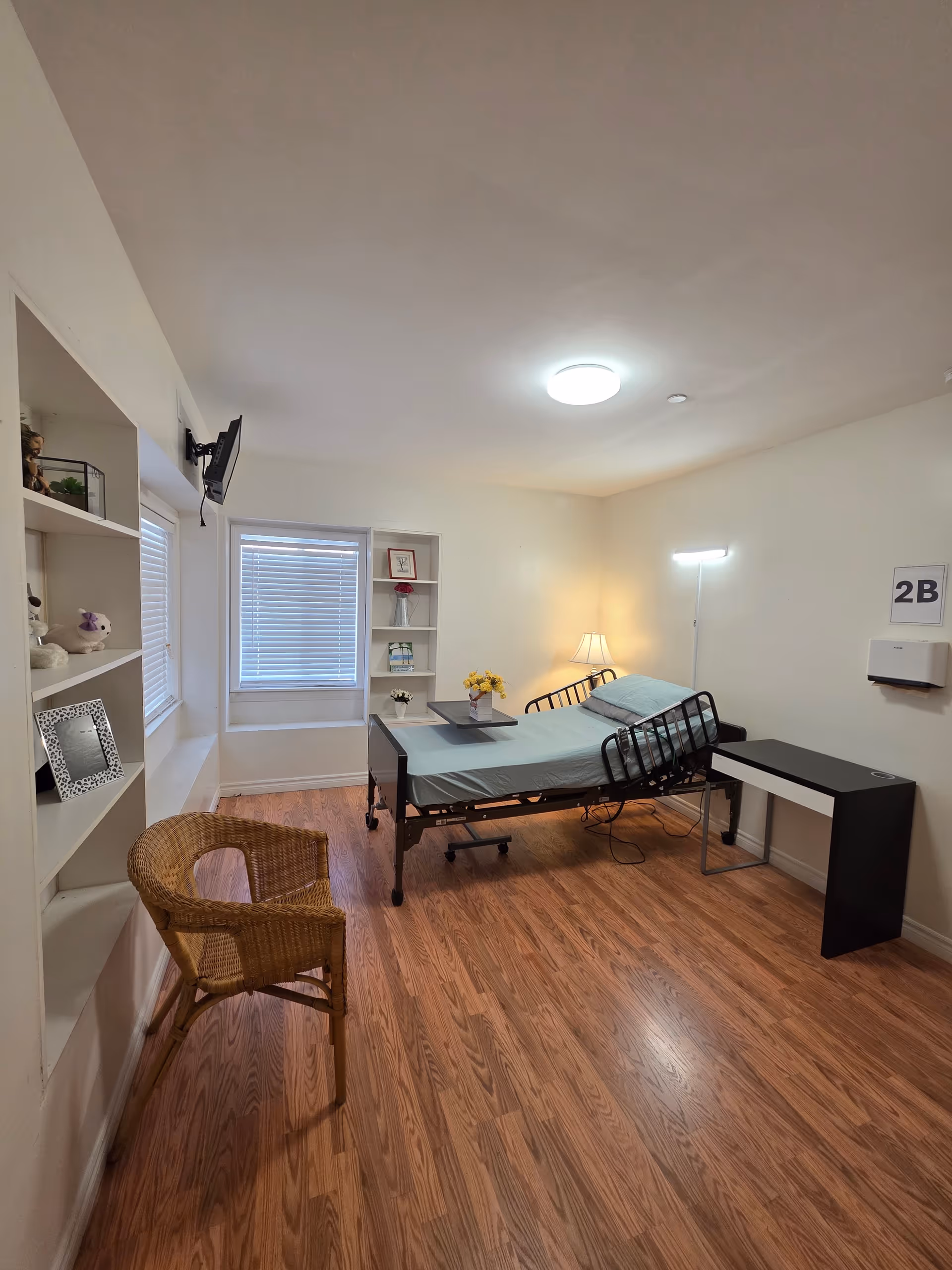 A small bedroom in a senior facility featuring an adjustable hospital bed, desk, and shelves.