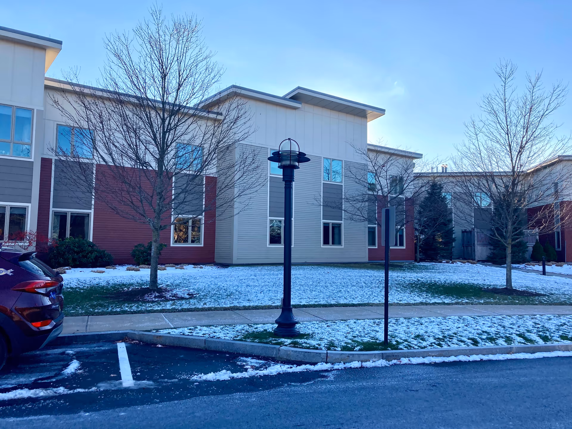 Front exterior of a two-story senior living building with bare trees, a lamp post, light snow on the lawn, and a parked car.