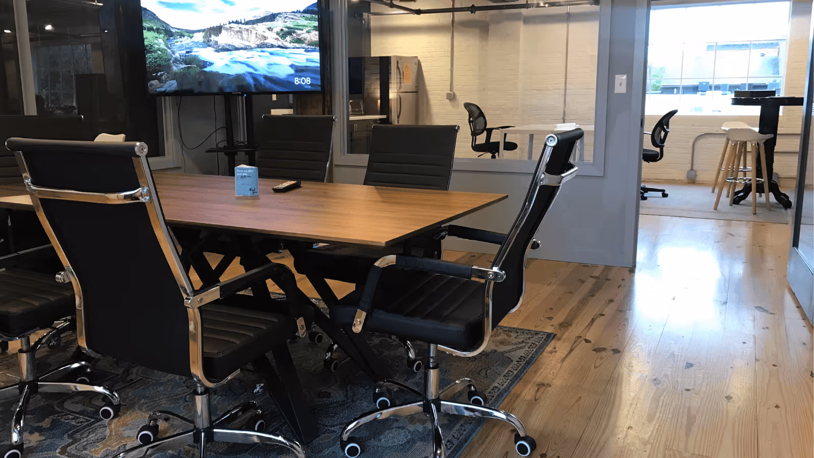 Indoor conference area with a wooden table, black chairs, TV, and glass-walled offices.