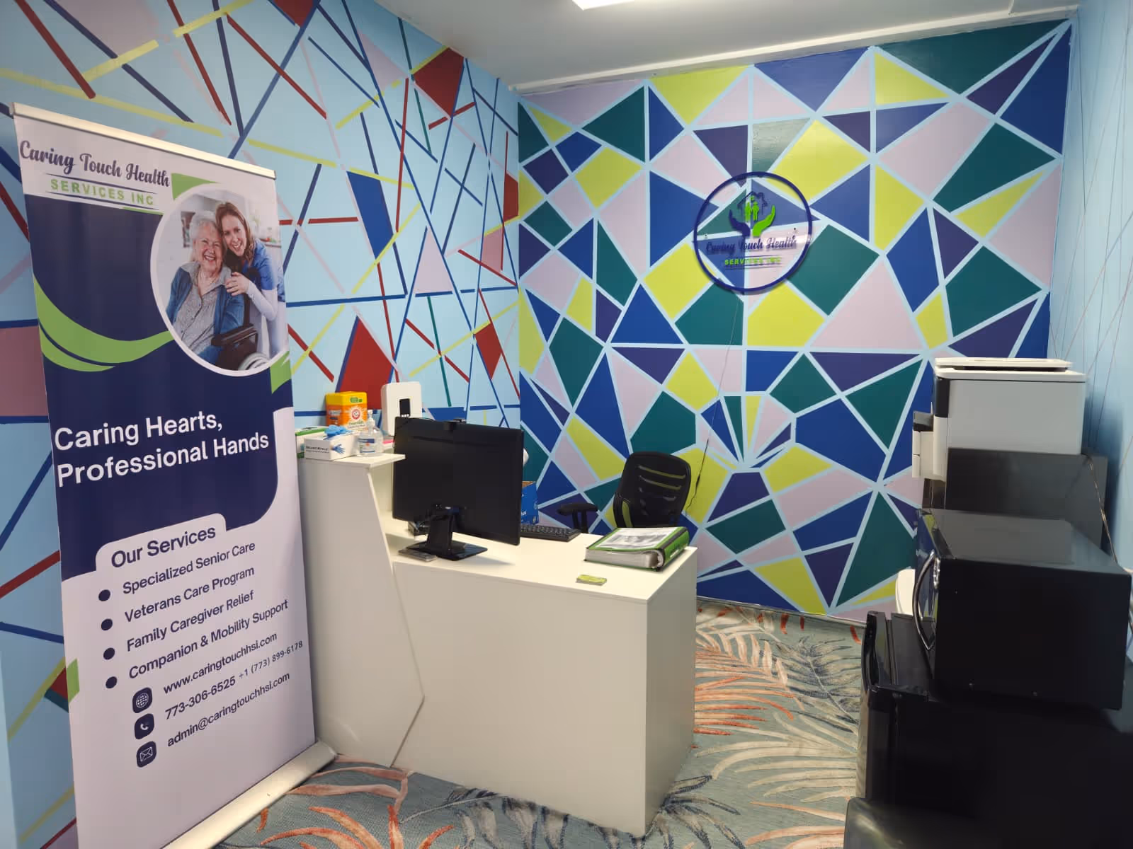 Reception area with colorful geometric mural walls, a white desk, and Caring Touch banner.