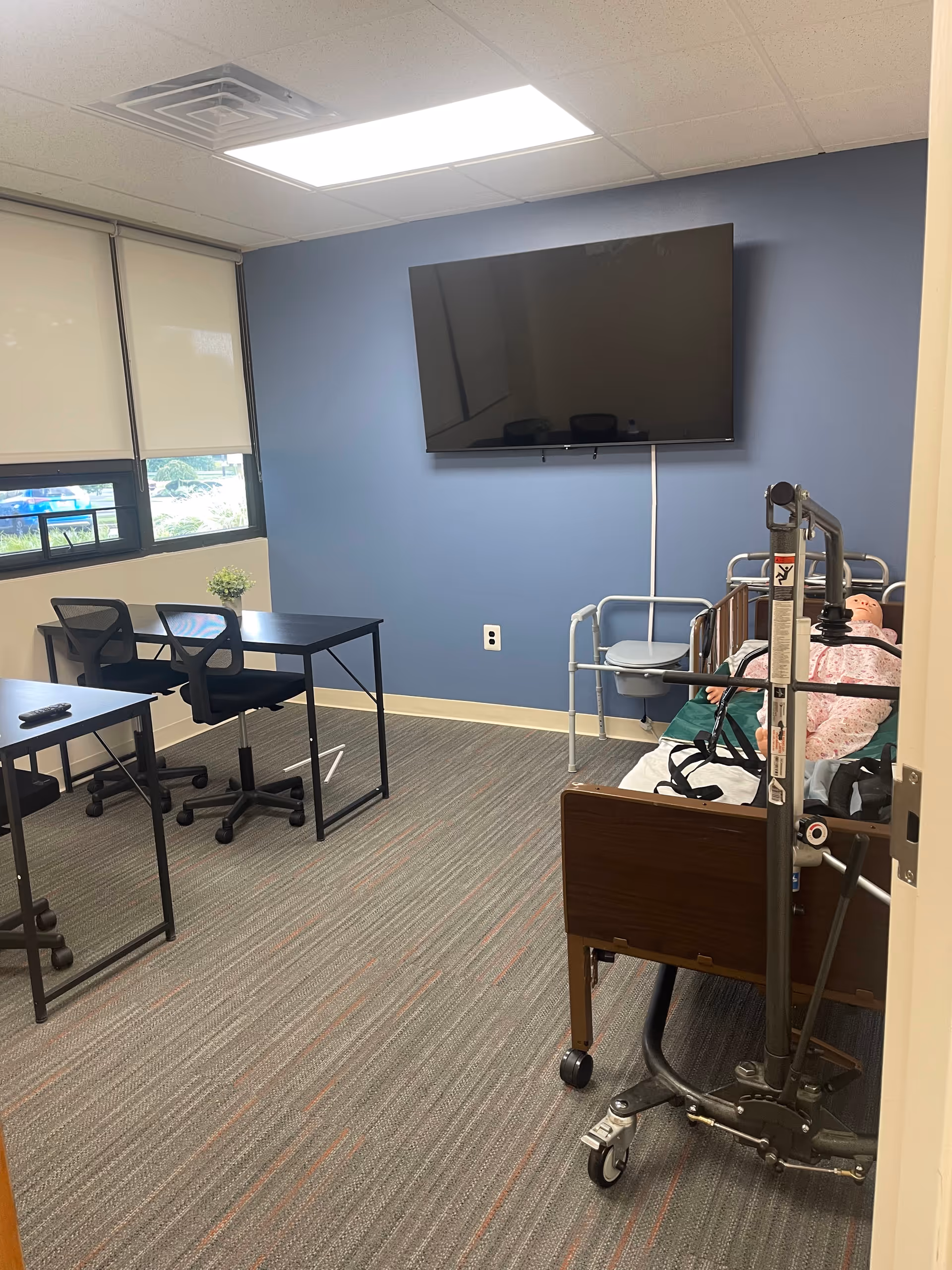 Patient room with a blue wall, mounted TV, desk, chairs, and an adjustable hospital bed.